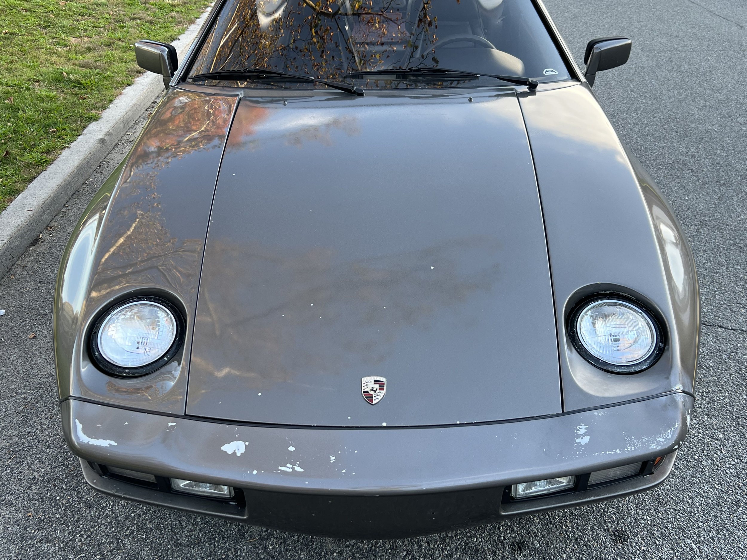 Front view of a gray Porsche sports car parked on the side of a road, with the Porsche emblem visible on the hood and reflections of trees on the windshield and hood.