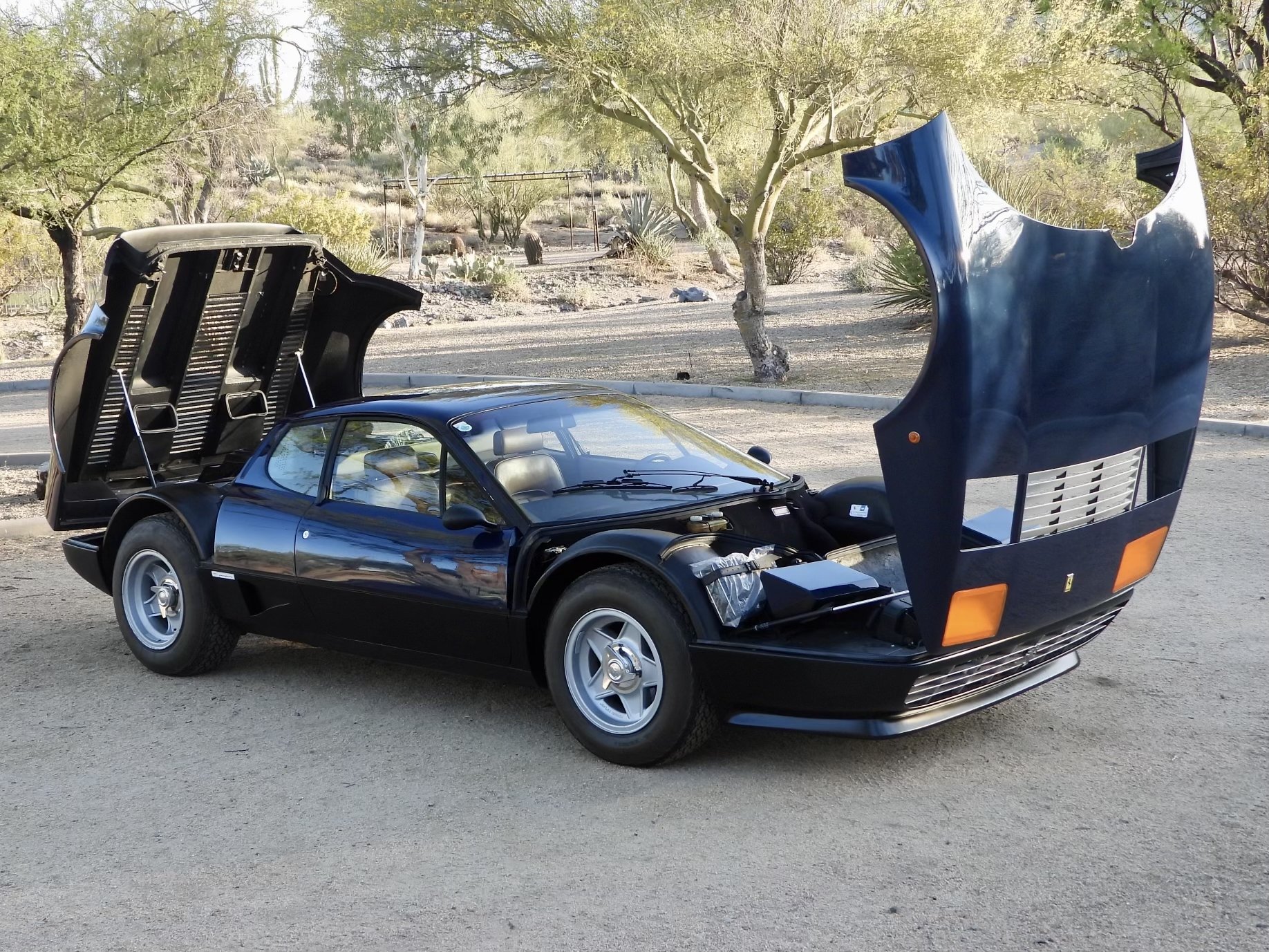 A Blu Dino Ferrari 512 BB sports car with its front and rear hoods open, parked on a desert landscape with trees and bushes in the background.