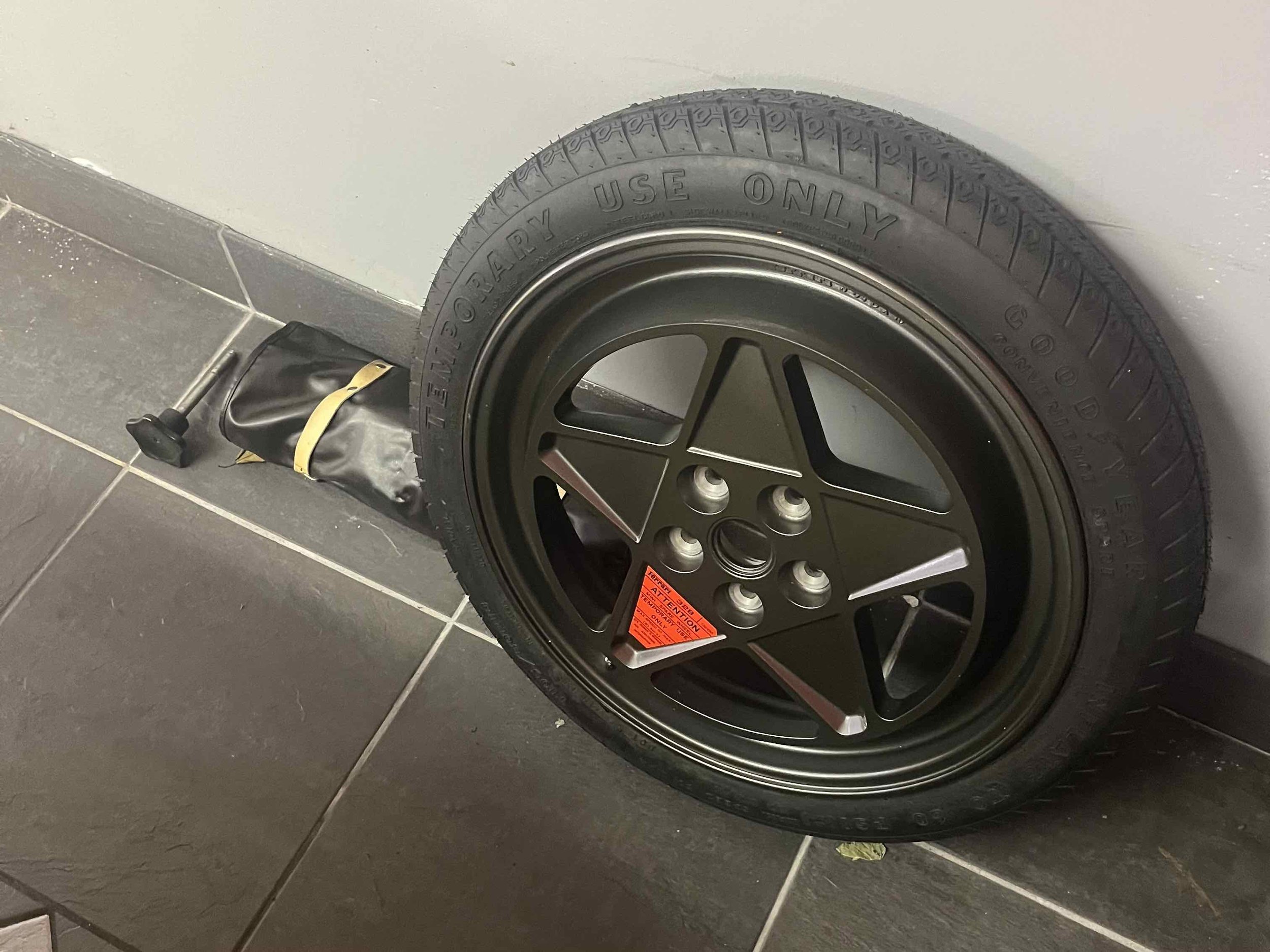 A wheel with a black rim and tire, and a black handle with a yellow strap attached to a small black bag, leaning against a gray wall on a black tiled floor.