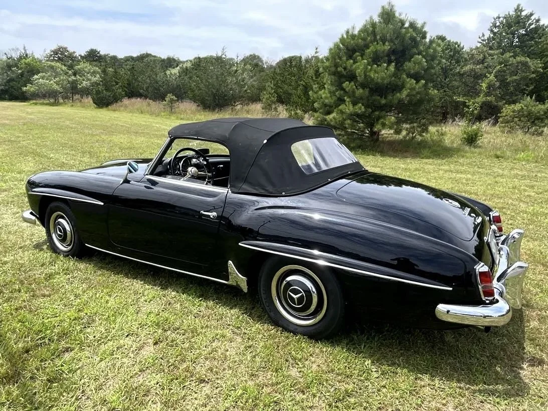 A vintage black convertible car with a soft top, parked on a grassy field with trees in the background.