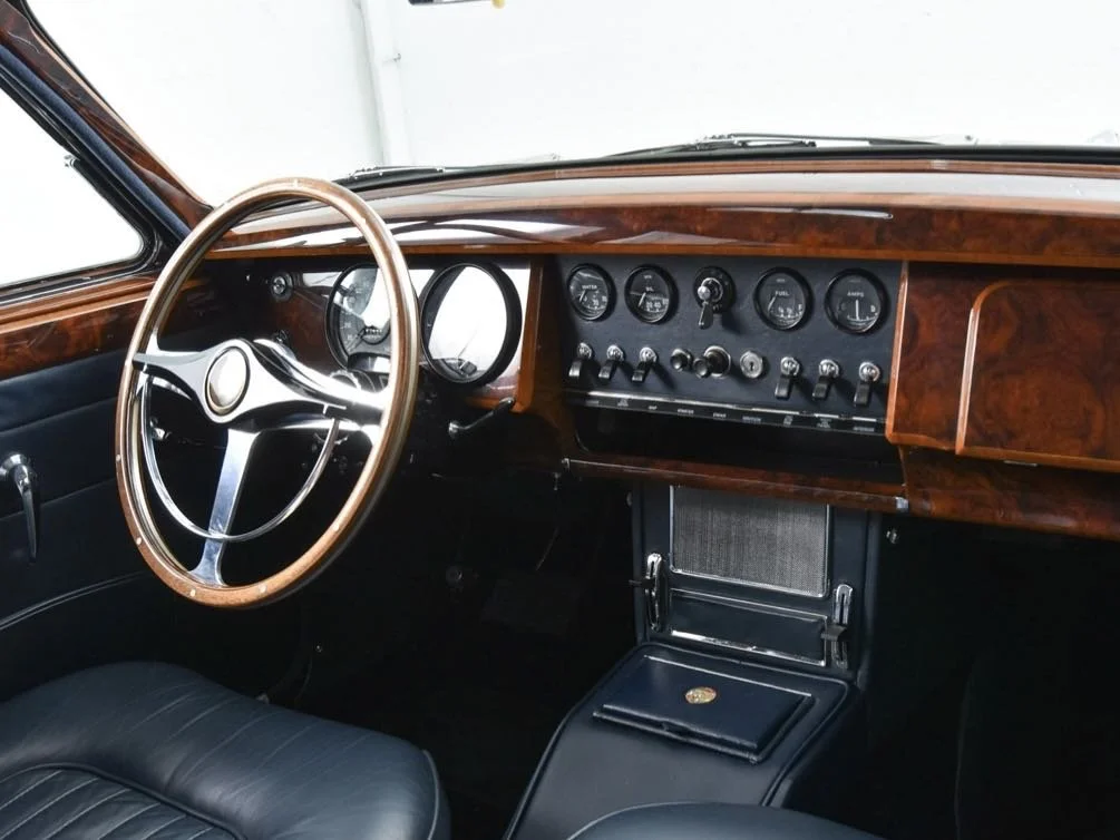 Interior of a vintage car with a wood dashboard, black steering wheel with wood trim, and black leather seats.
