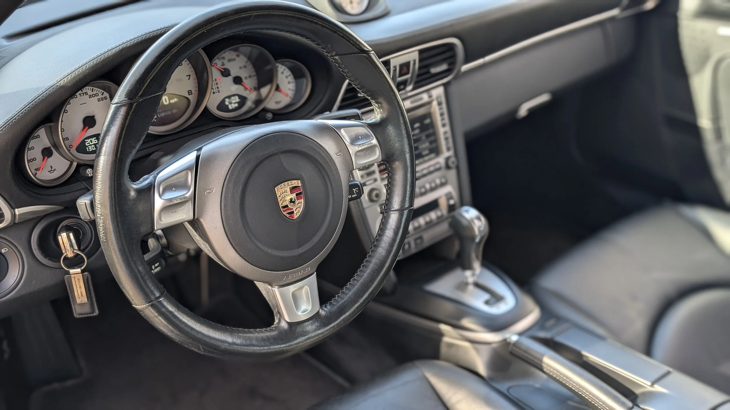 Interior of a car dashboard showing a steering wheel with a Porsche logo, speedometer, tachometer, and gear shift lever.