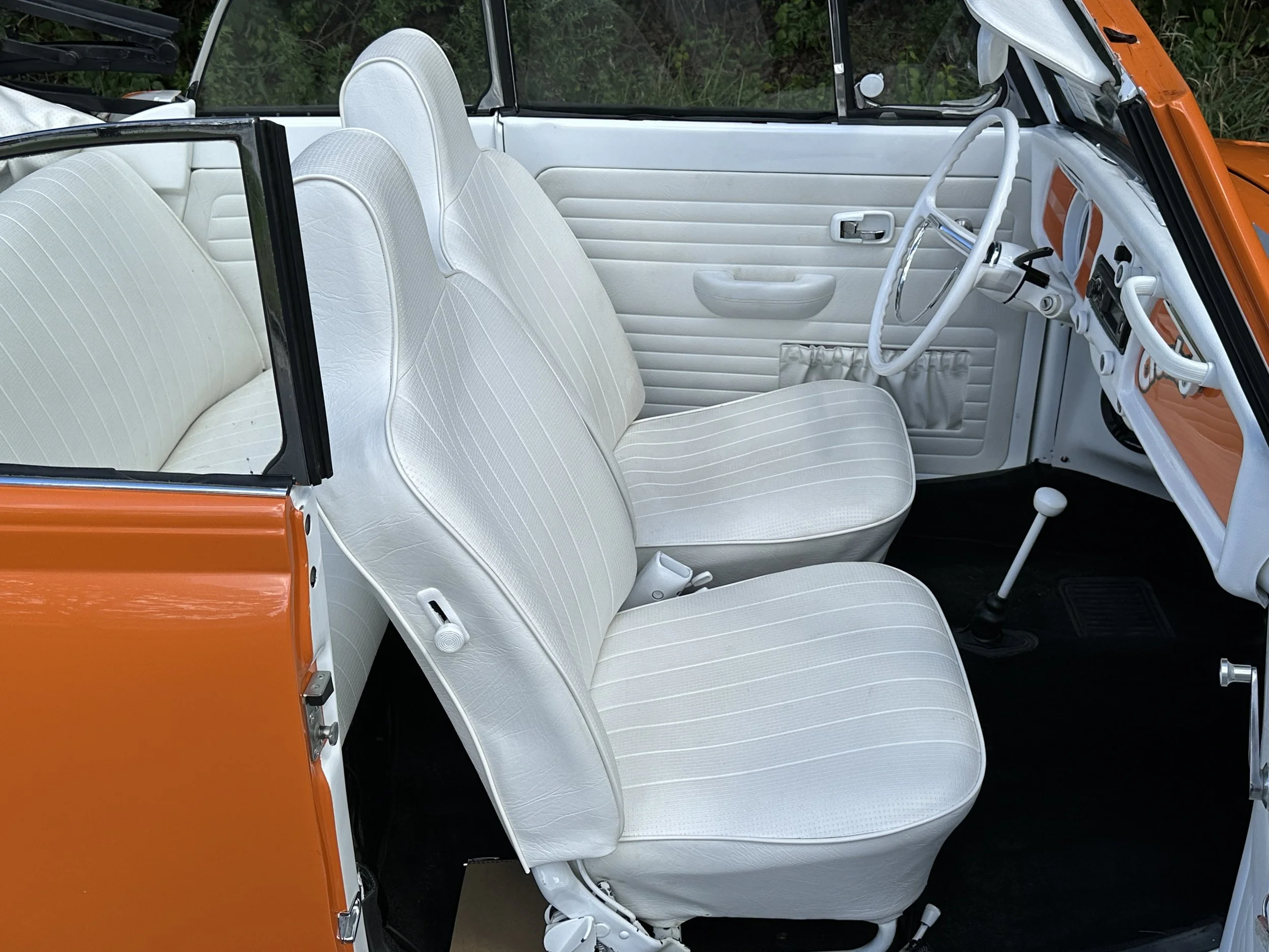 Interior of a vintage orange and white car with white seats, white steering wheel, and dashboard, viewed from the open door.