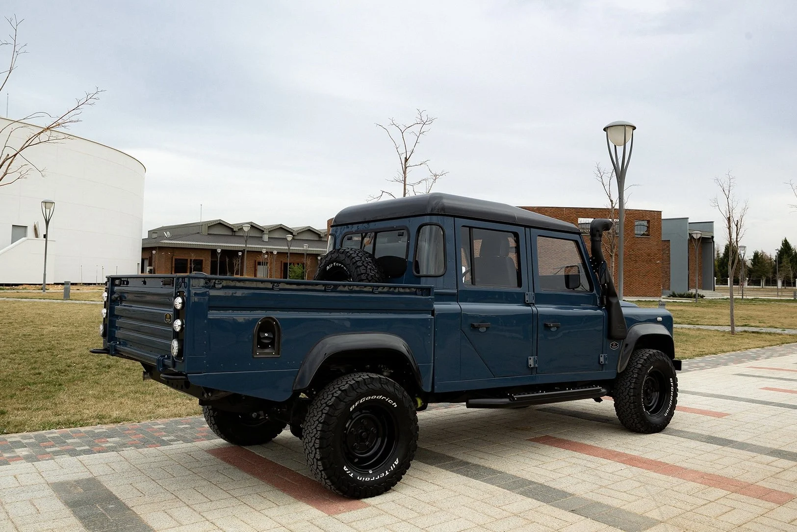 A vintage blue pickup truck with a black roof and off-road tires parked on a paved walkway in an outdoor park with grass, leafless trees, modern buildings, and street lamps in the background.