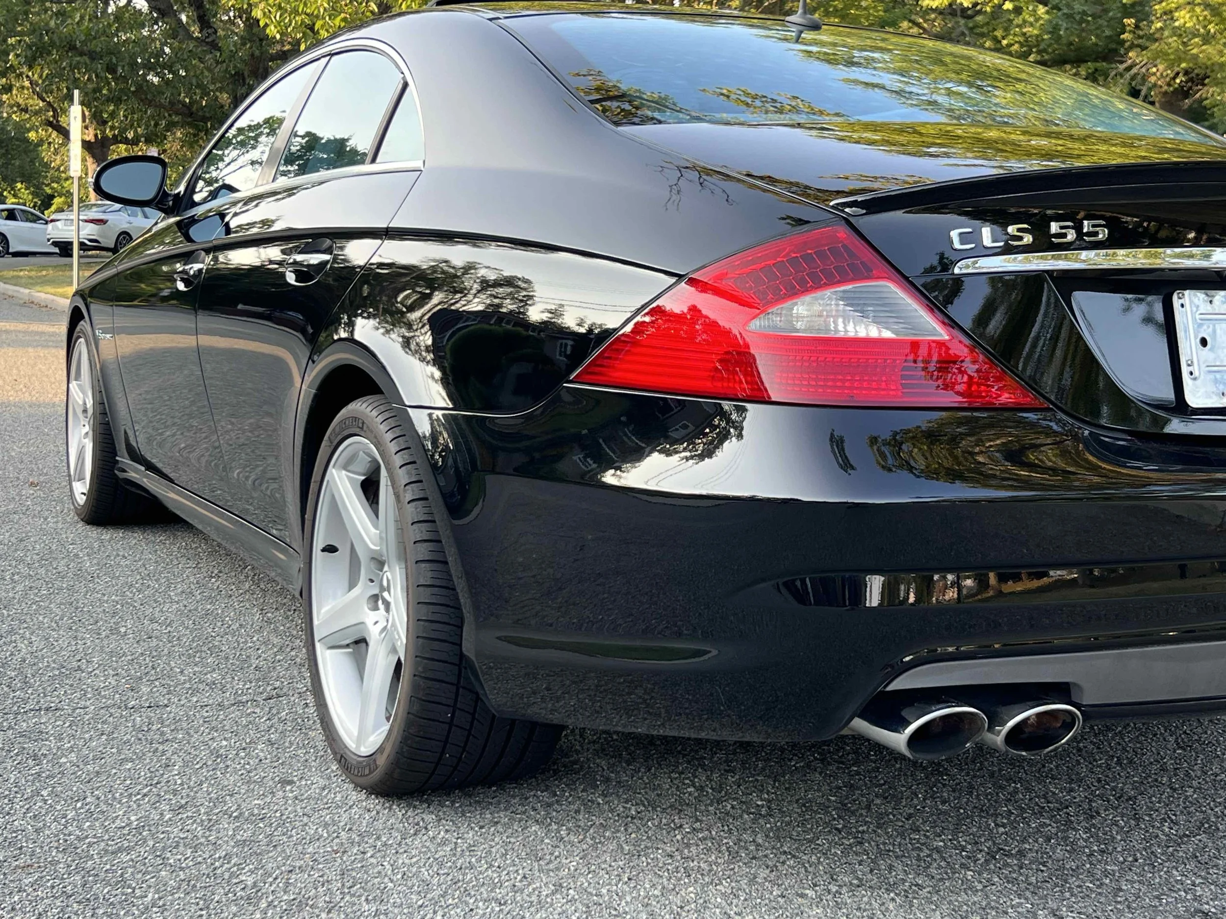 Black Mercedes-Benz CLS 55 with silver wheels and dual exhaust pipes parked on the street, with trees and other cars in the background.