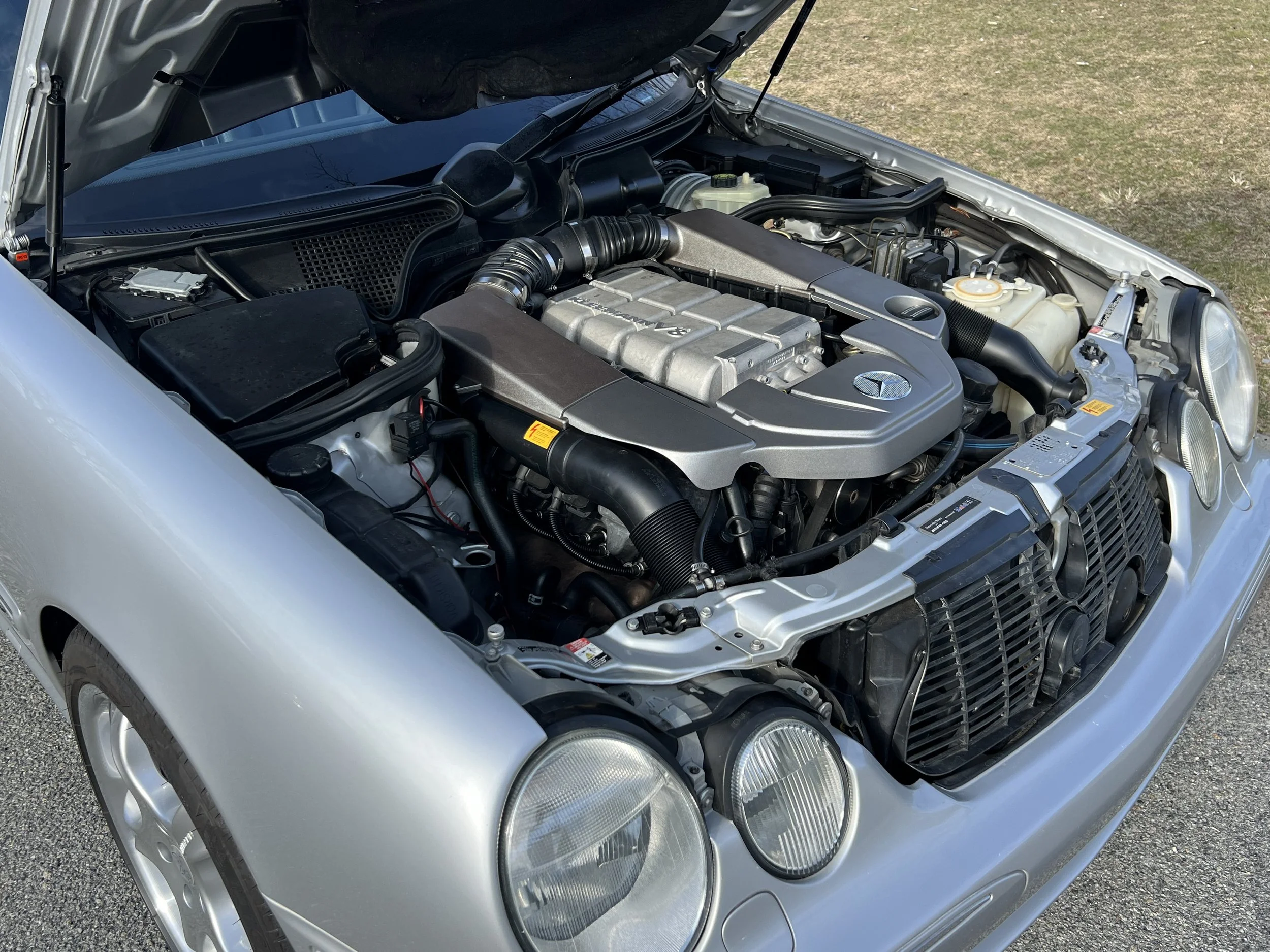 A silver car with the hood open, showing the engine and various engine components under a clear sky and on a paved surface with grass nearby.