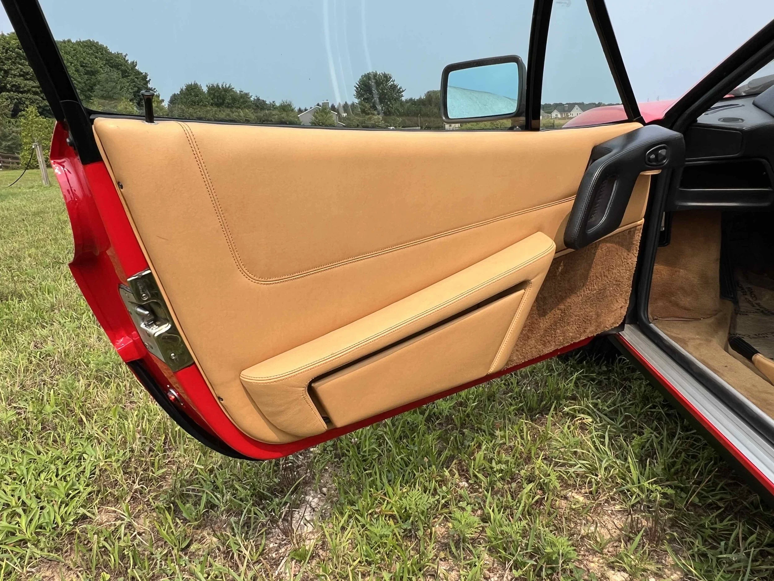 The interior door panel of a red vintage car with tan leather and carpet accents, parked on grass with trees and a few houses in the background.