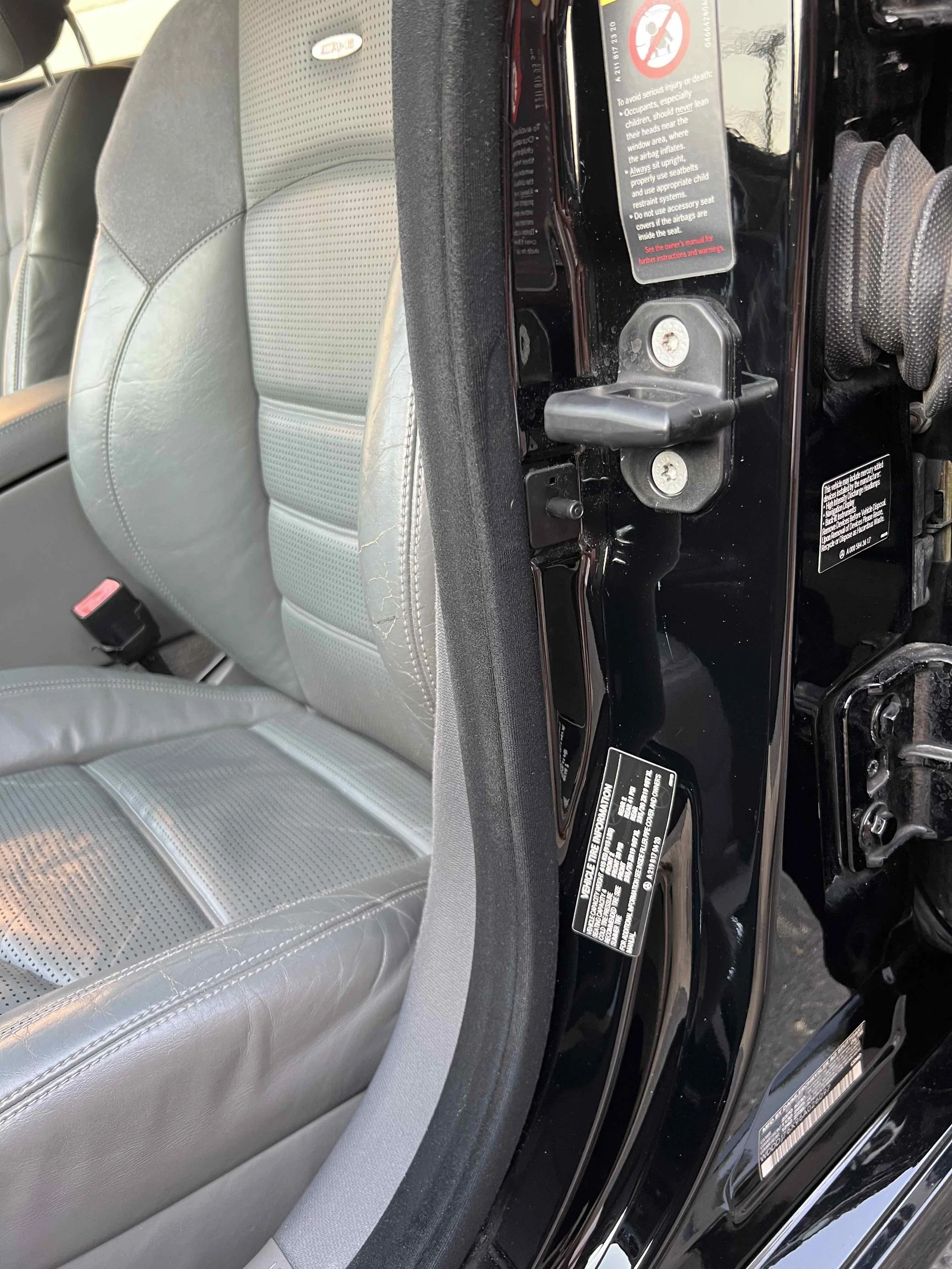 Close-up of a gray leather car seat and part of a black door frame with safety stickers and a door latch.