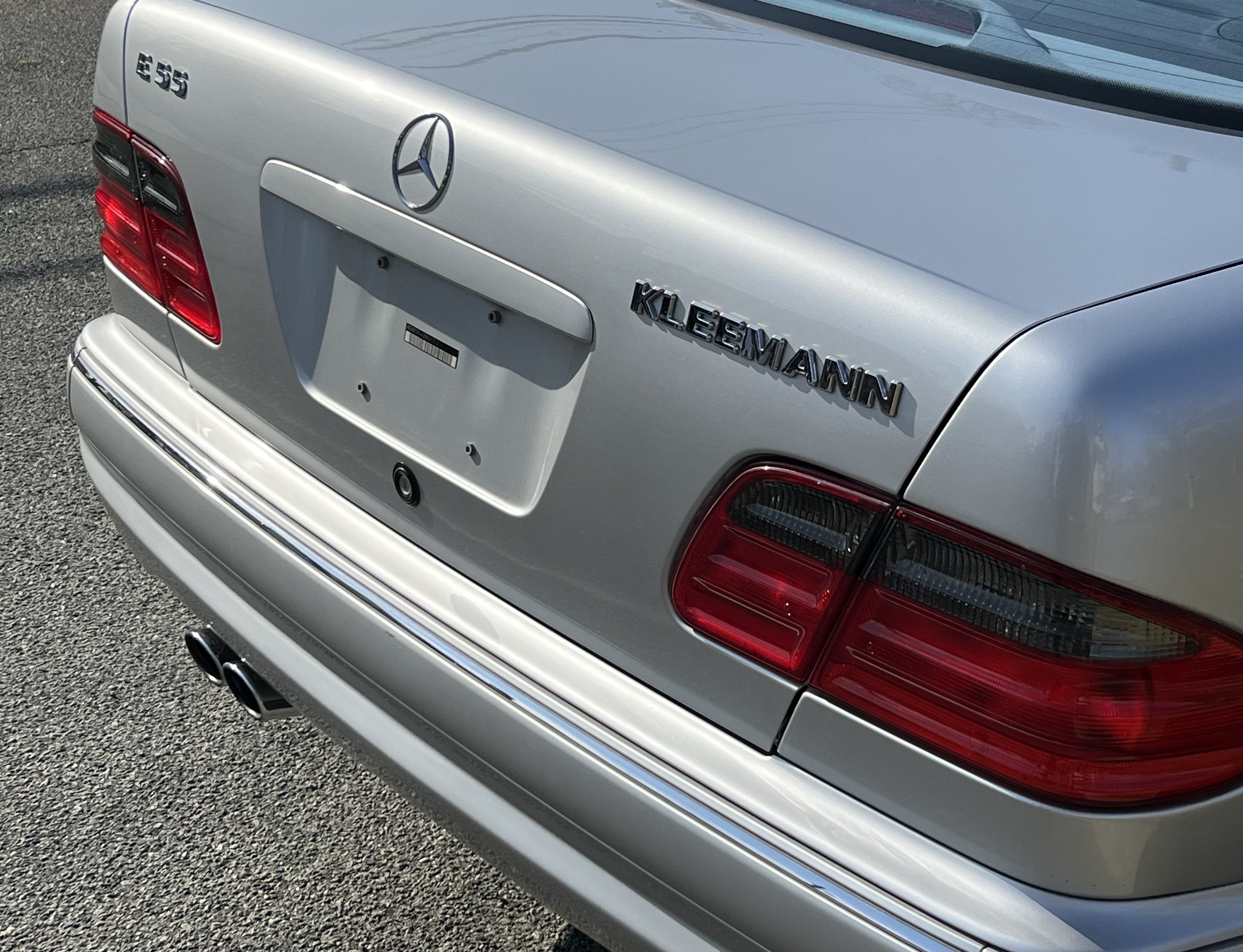 The rear of a silver Mercedes-Benz E55 AMG car displaying the Mercedes emblem, model badge, tail lights, and dual exhaust pipes.