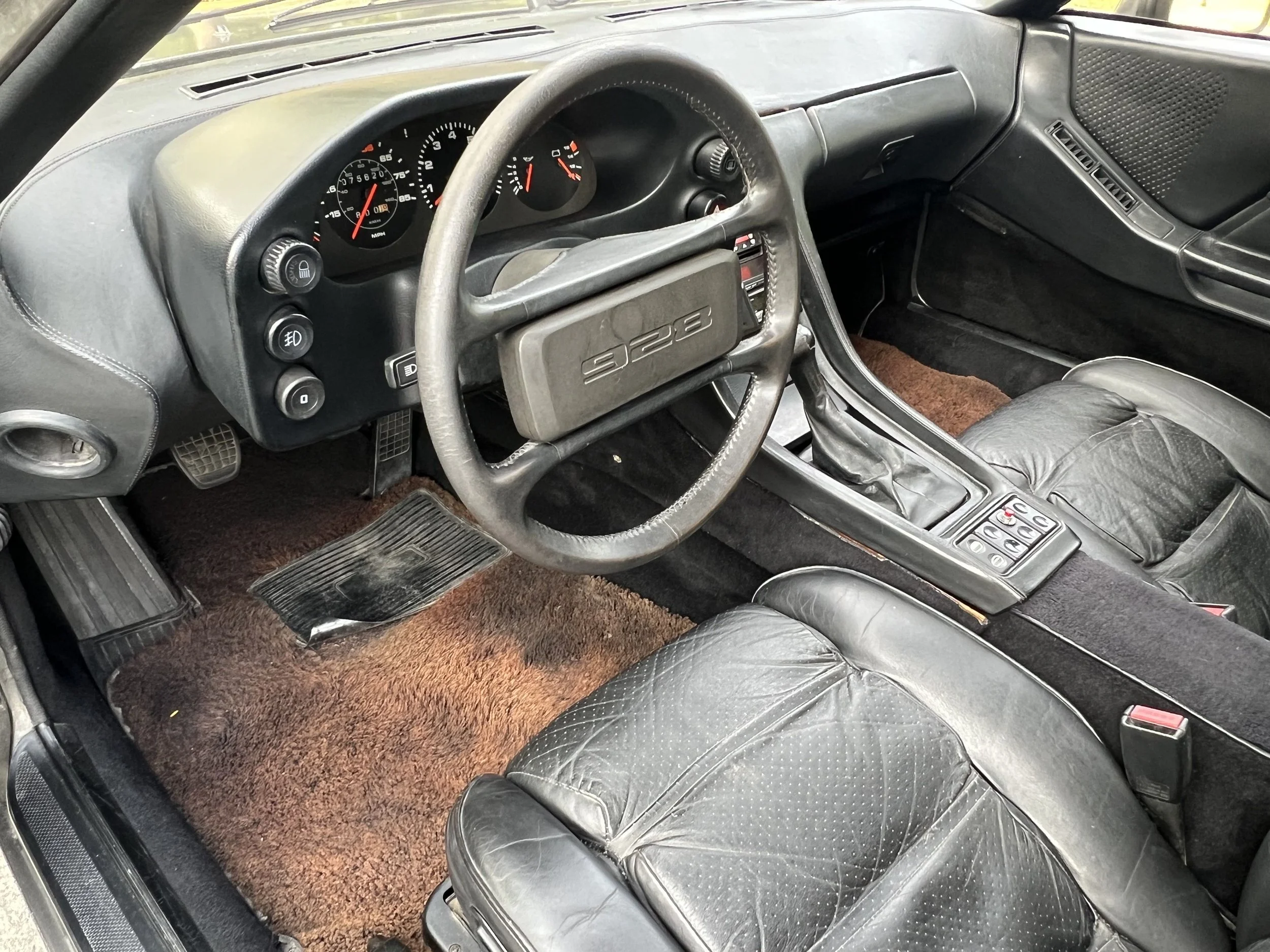 Interior of a vintage black Porsche 928 with black leather seats, a brown carpet floor, and a dashboard with analog gauges and control buttons.