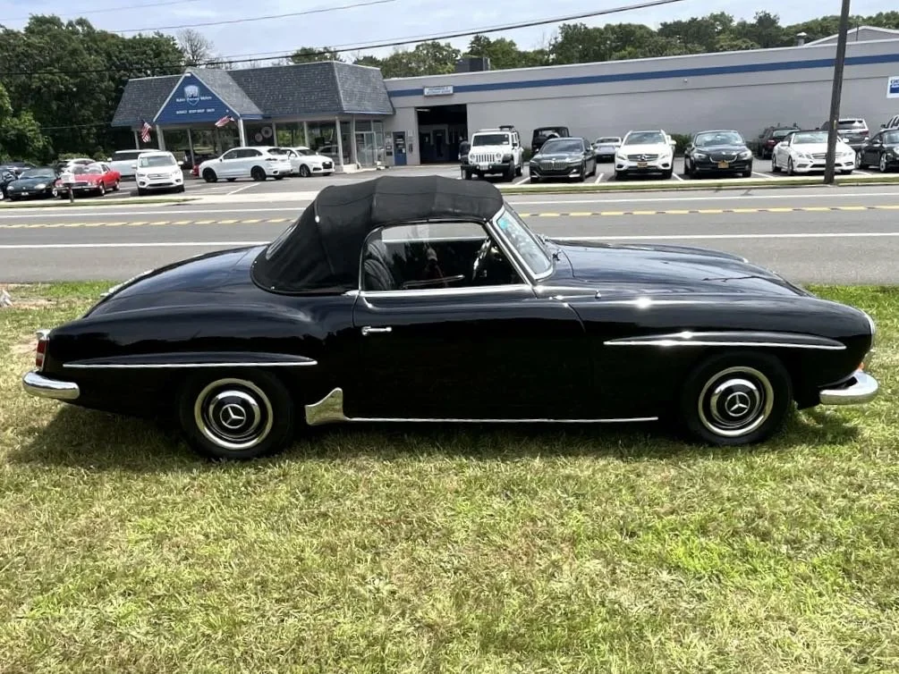Black vintage convertible car parked on grass next to a busy street with a car dealership in the background.