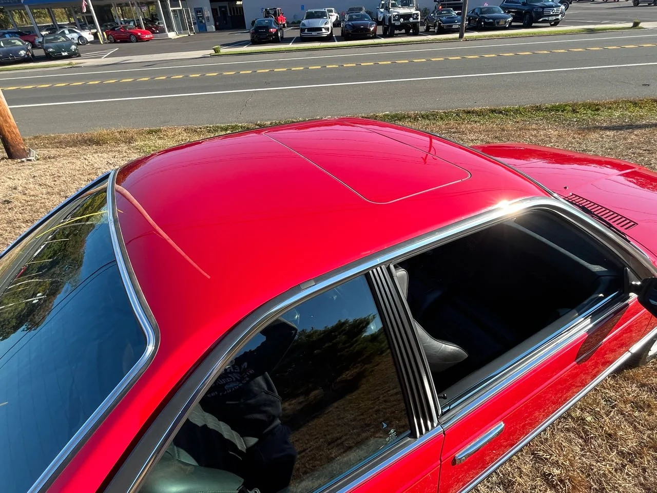 Red vintage car parked on grass with a reflection of trees on the windshield, and a street with parked cars and a shopping area in the background.