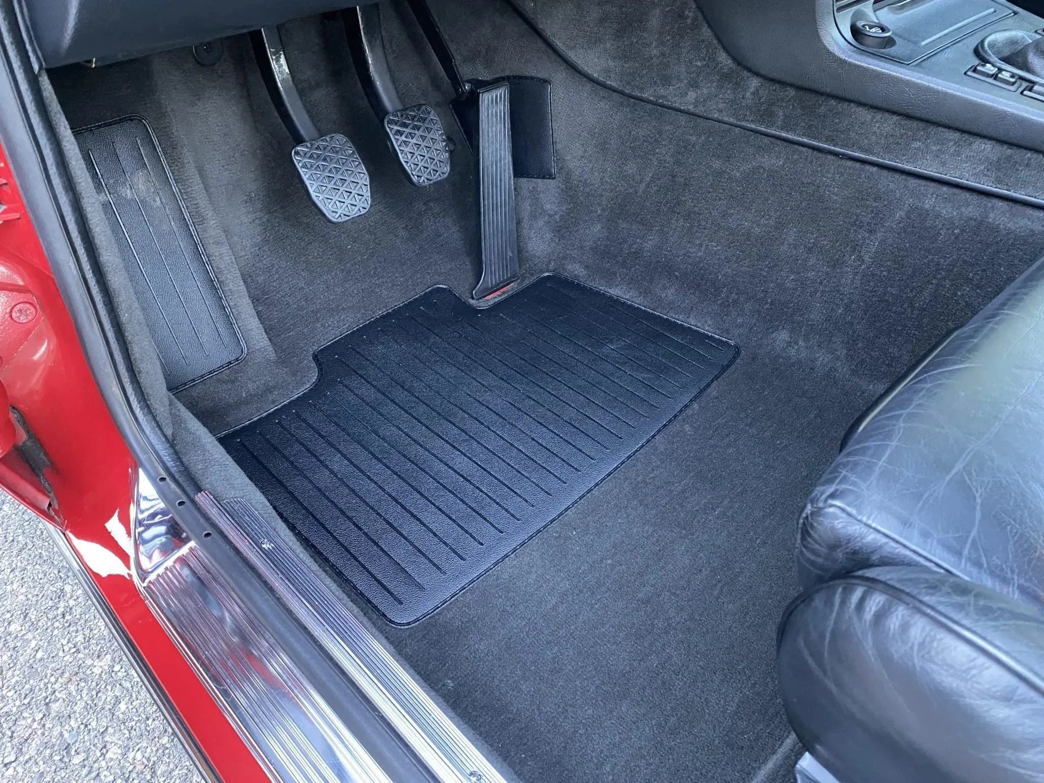 The image shows the driver's side footwell of a car, with three pedals (clutch, brake, accelerator), a black rubber floor mat, and part of a black leather seat on the right.