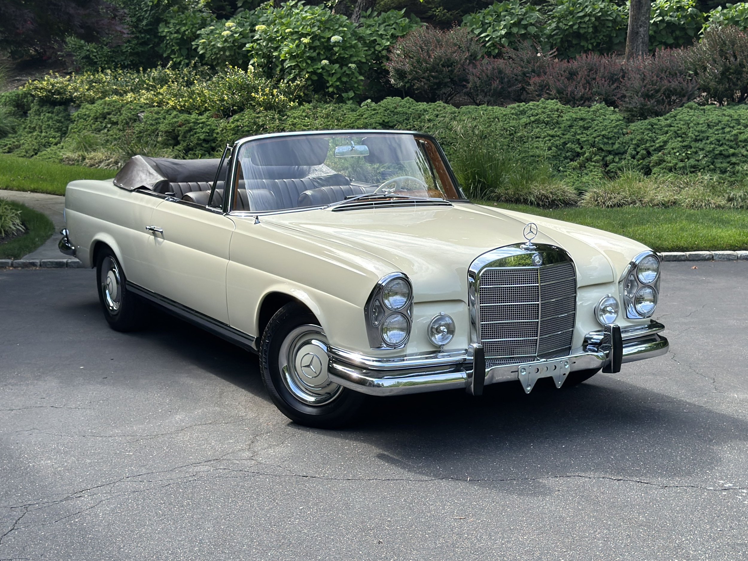 A vintage cream-colored Mercedes-Benz convertible car parked on a paved area with a green landscaped background.