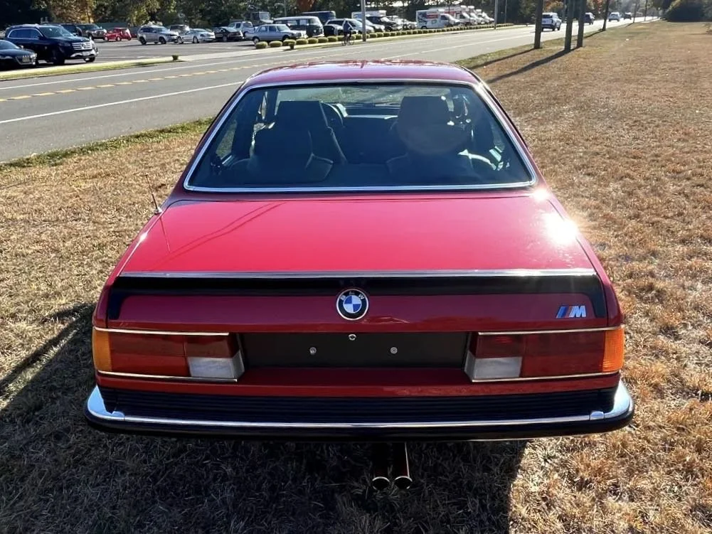 Red classic BMW M car parked on grass near a road, with a row of cars in the background.