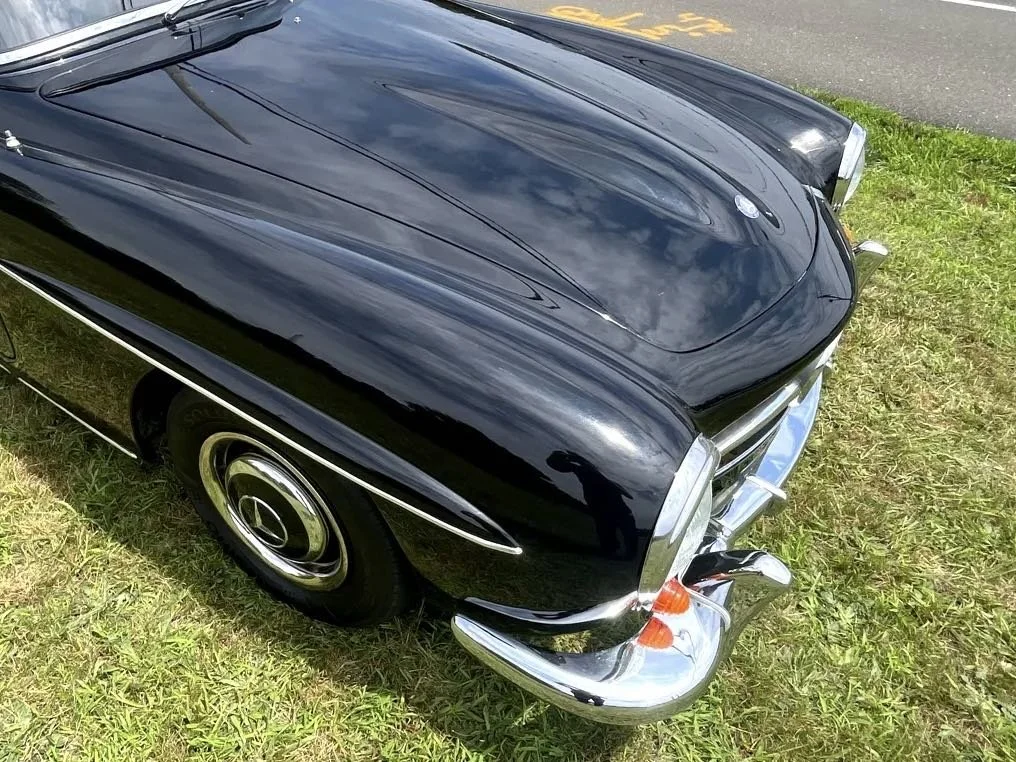 A vintage black Mercedes-Benz car parked on grass next to a paved road, with reflections of clouds visible on its hood.