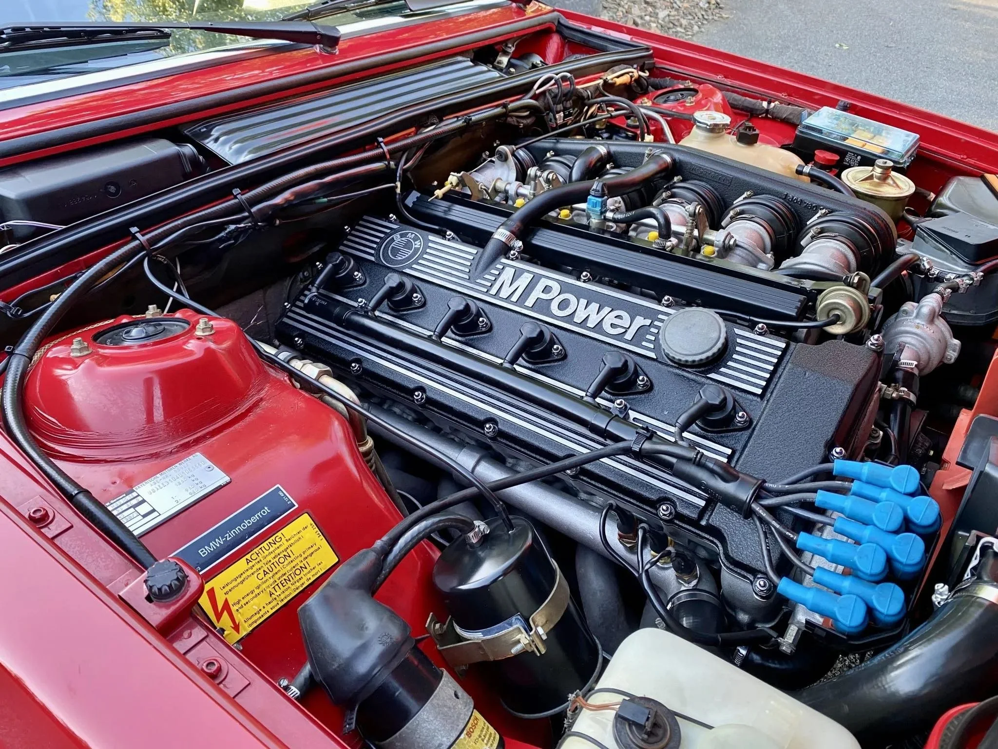 Under the hood of a red BMW car, showing the engine with an 'M Power' label on the valve cover, various engine components, hoses, and blue spark plug wires.