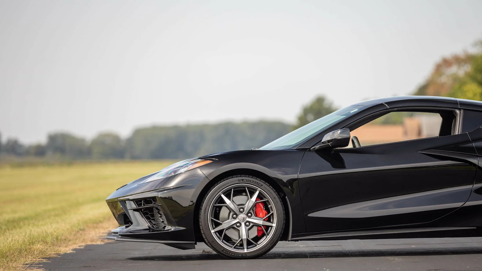Black sports car parked on a paved road with fields and trees in the background during daytime.