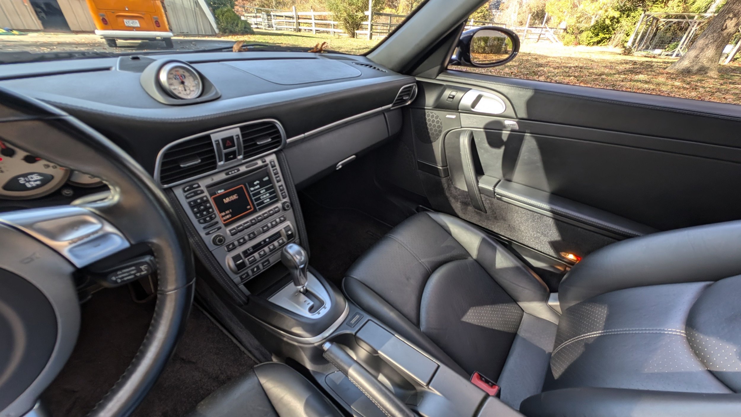 Interior of a sports car showing the dashboard, center console with control buttons, automatic gear shift, and black leather seats, with a view of the road and trees outside.