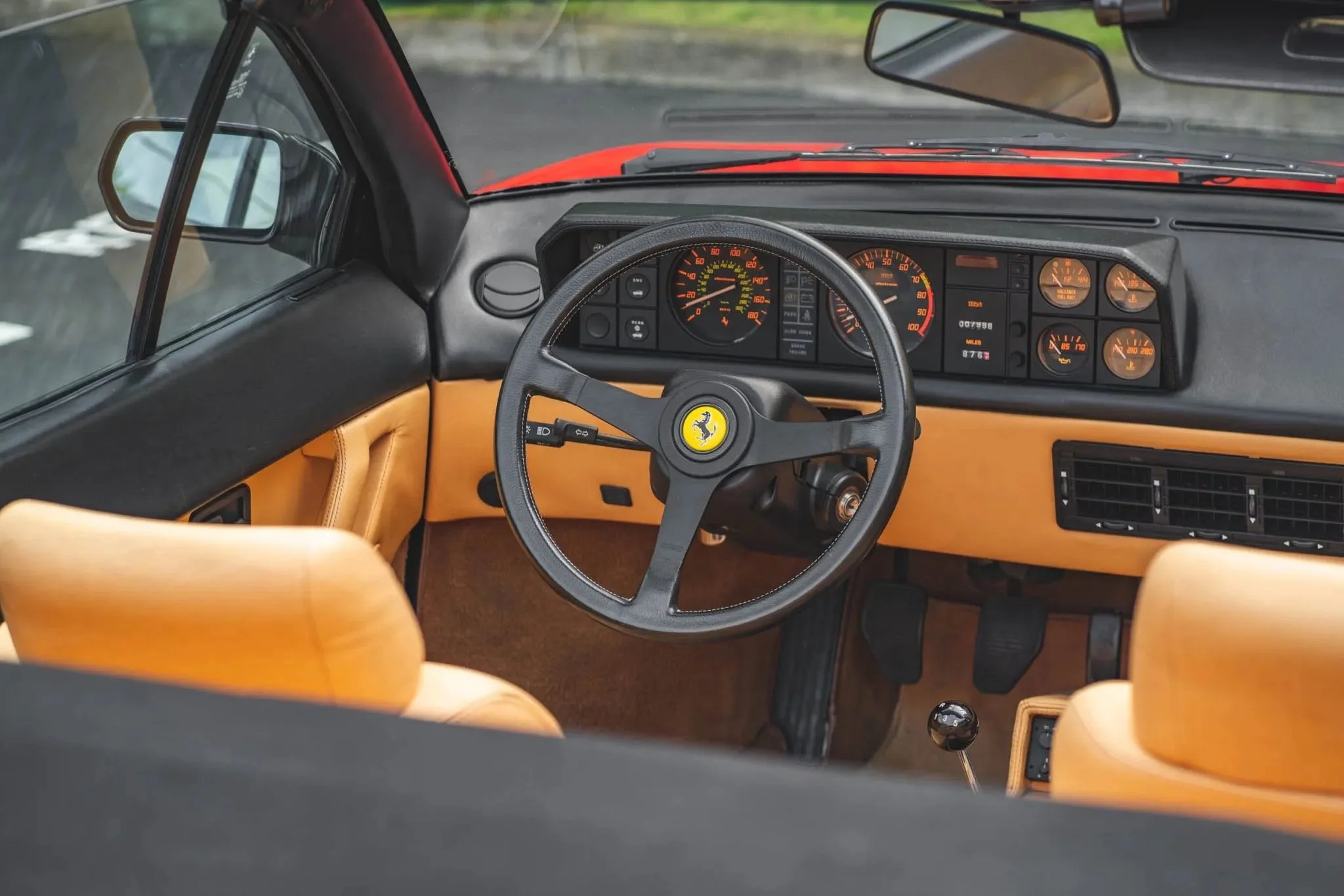 Interior of a vintage black and tan Ferrari sports car showing the dashboard, steering wheel with Ferrari logo, brown seats, and instrument panel with gauges.