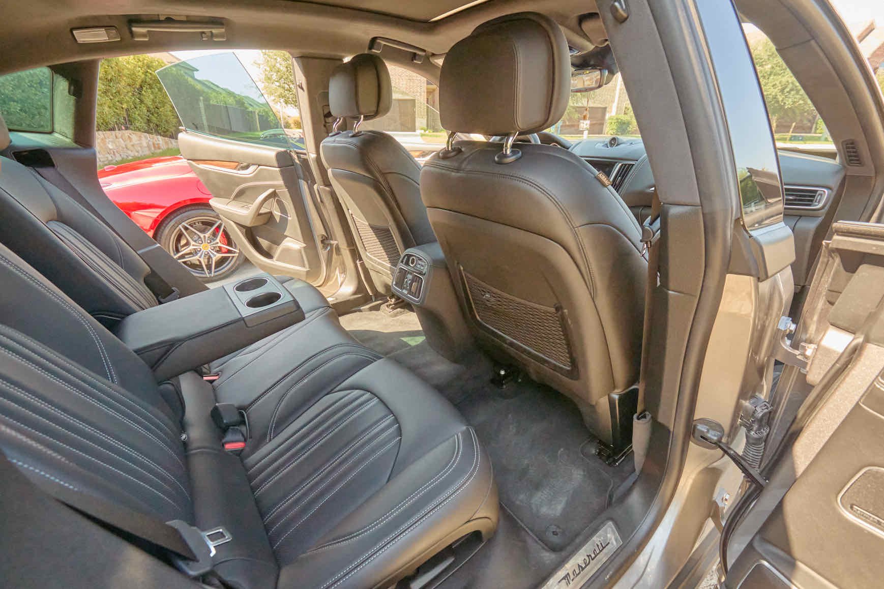The interior of a luxury car showing black leather seats, with the backseat area, rear of the front seats, and center console visible. A red sports car is partially outside, seen through the open rear side door.