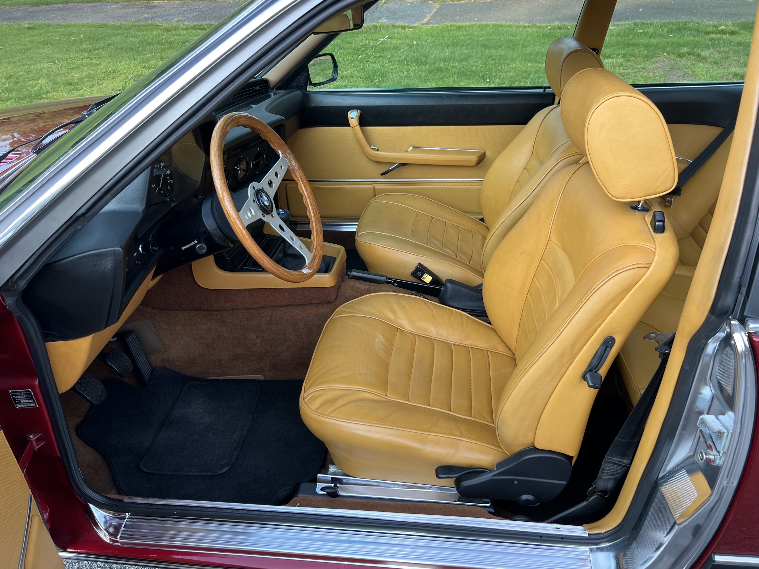 Interior of a vintage BMW 630 csi car with tan leather seats, wooden steering wheel, black dashboard, and black floor mats.