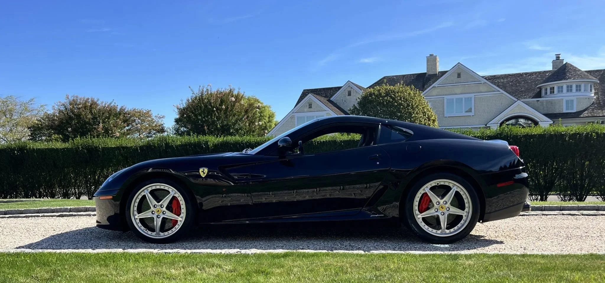 A sleek black Ferrari sports car parked on a driveway in front of a hedge and a large house with a clear blue sky overhead.