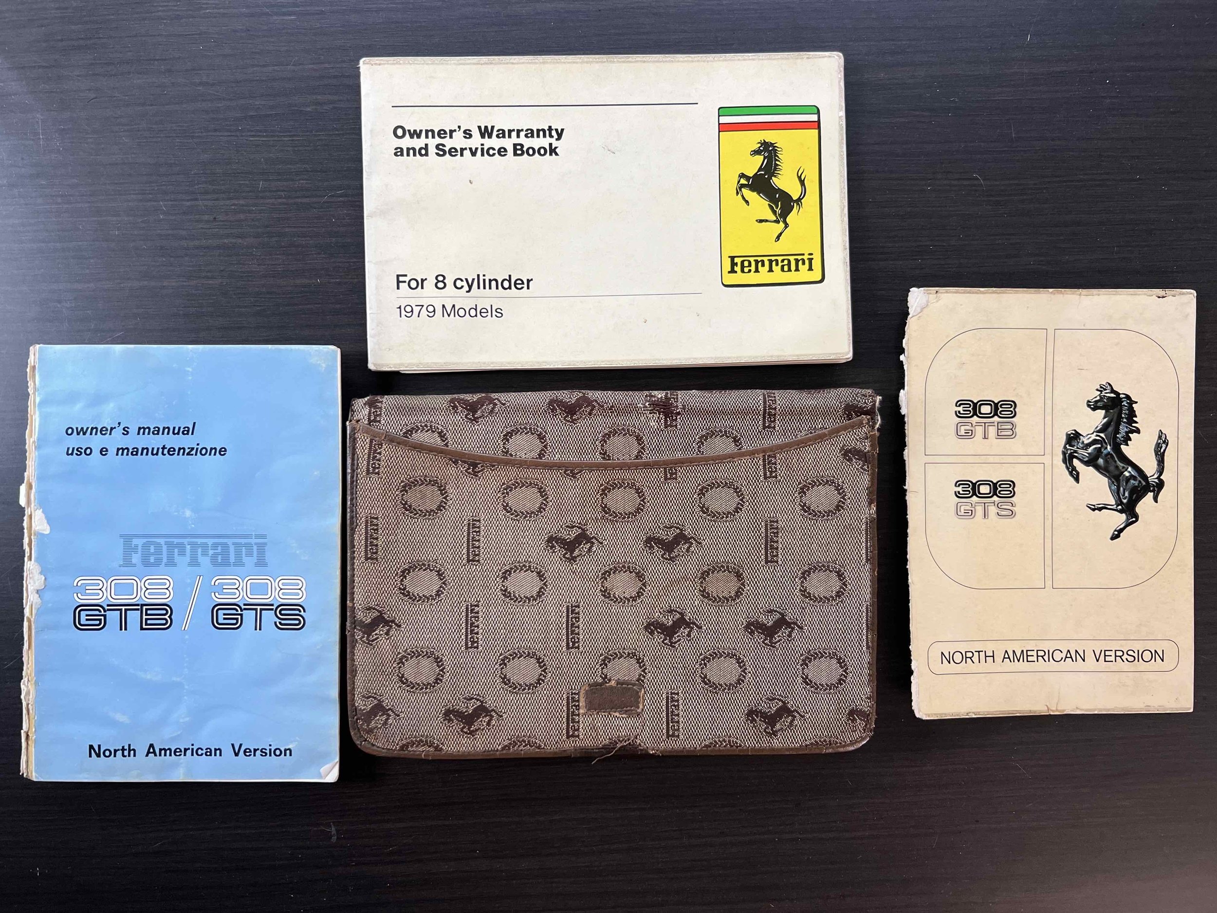 Collection of vintage Ferrari owner's manuals, warranty booklets, and a brown patterned folder with Ferrari logos, all arranged on a dark surface.