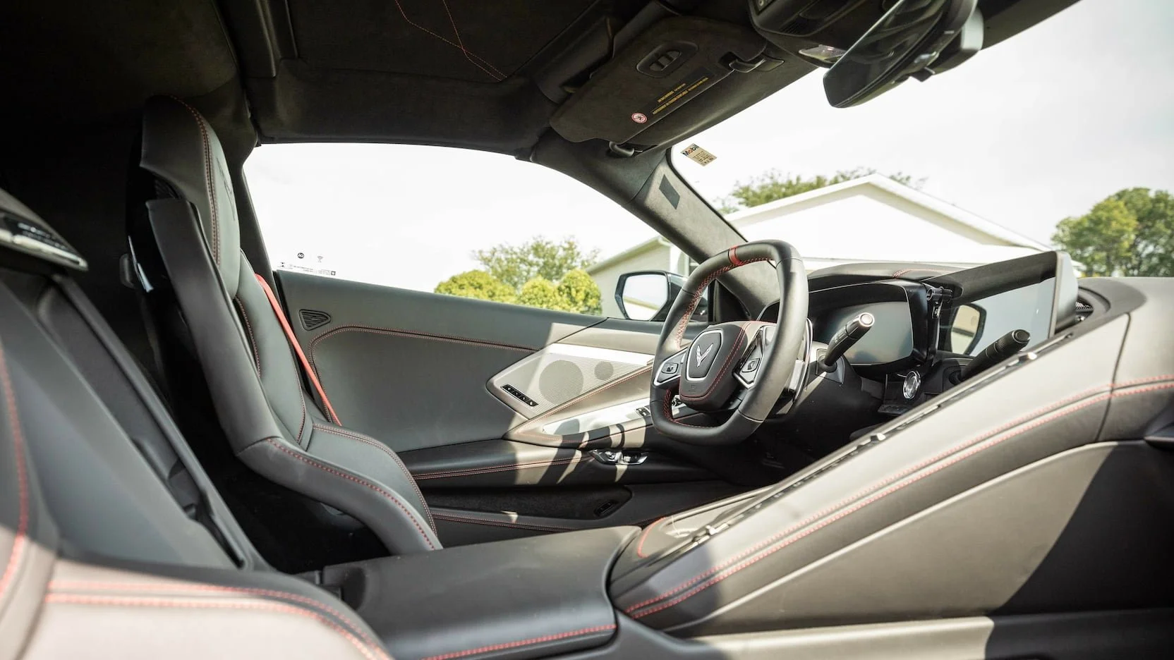 The interior of a modern sports car showing the driver's seat, steering wheel, dashboard, and door panel with red stitching details, outside view of a house and trees.