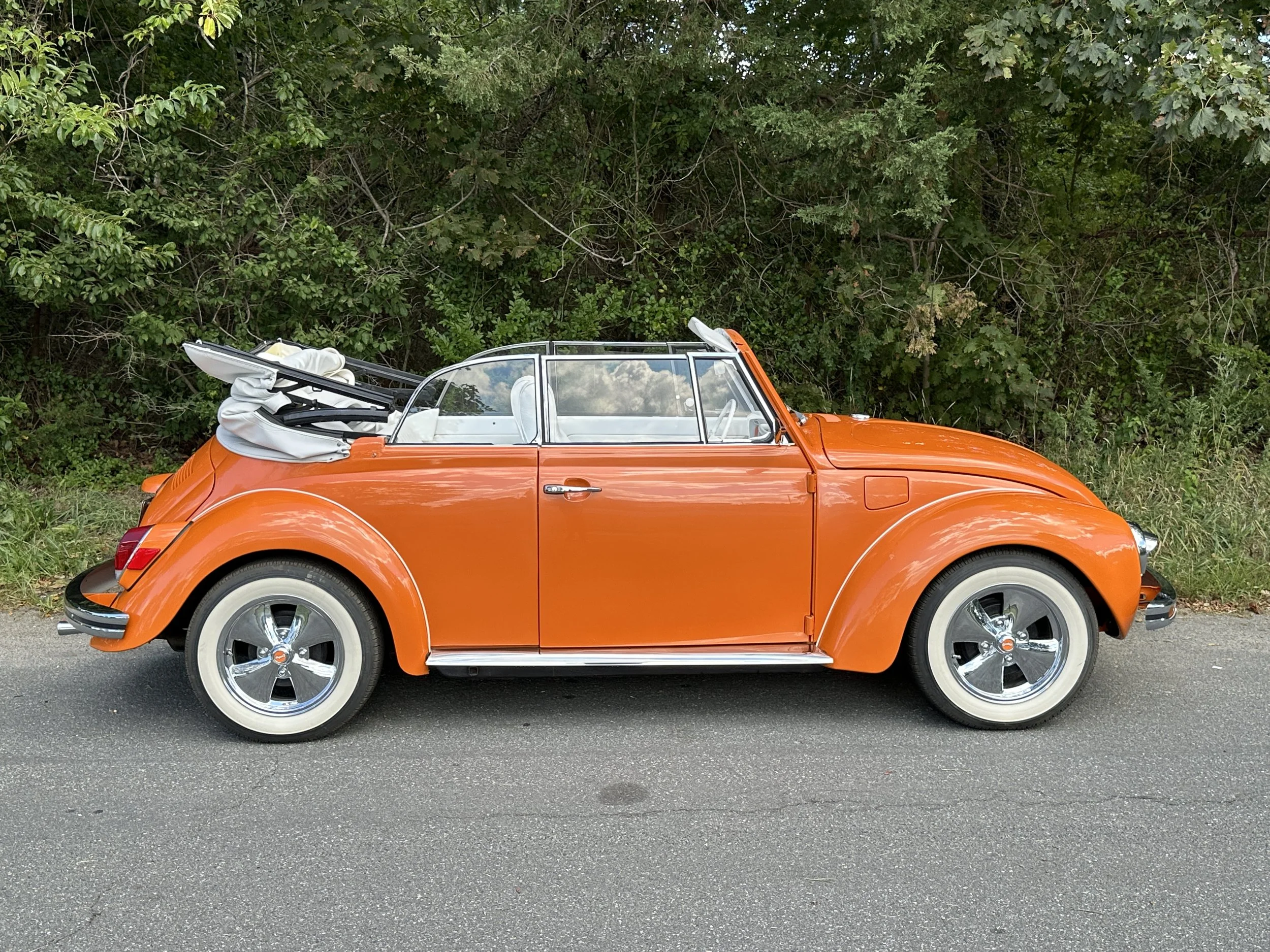 Orange vintage Volkswagen Beetle with a folded white soft top, parked on a road with green trees and bushes in the background.