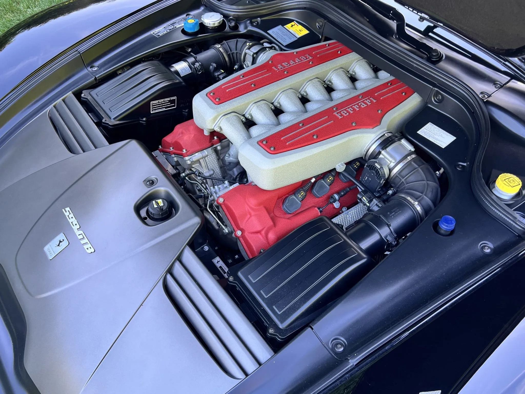 The image shows the engine bay of a Ferrari sports car, with a prominently displayed red and silver Ferrari engine cover, various engine components, hoses, and parts.