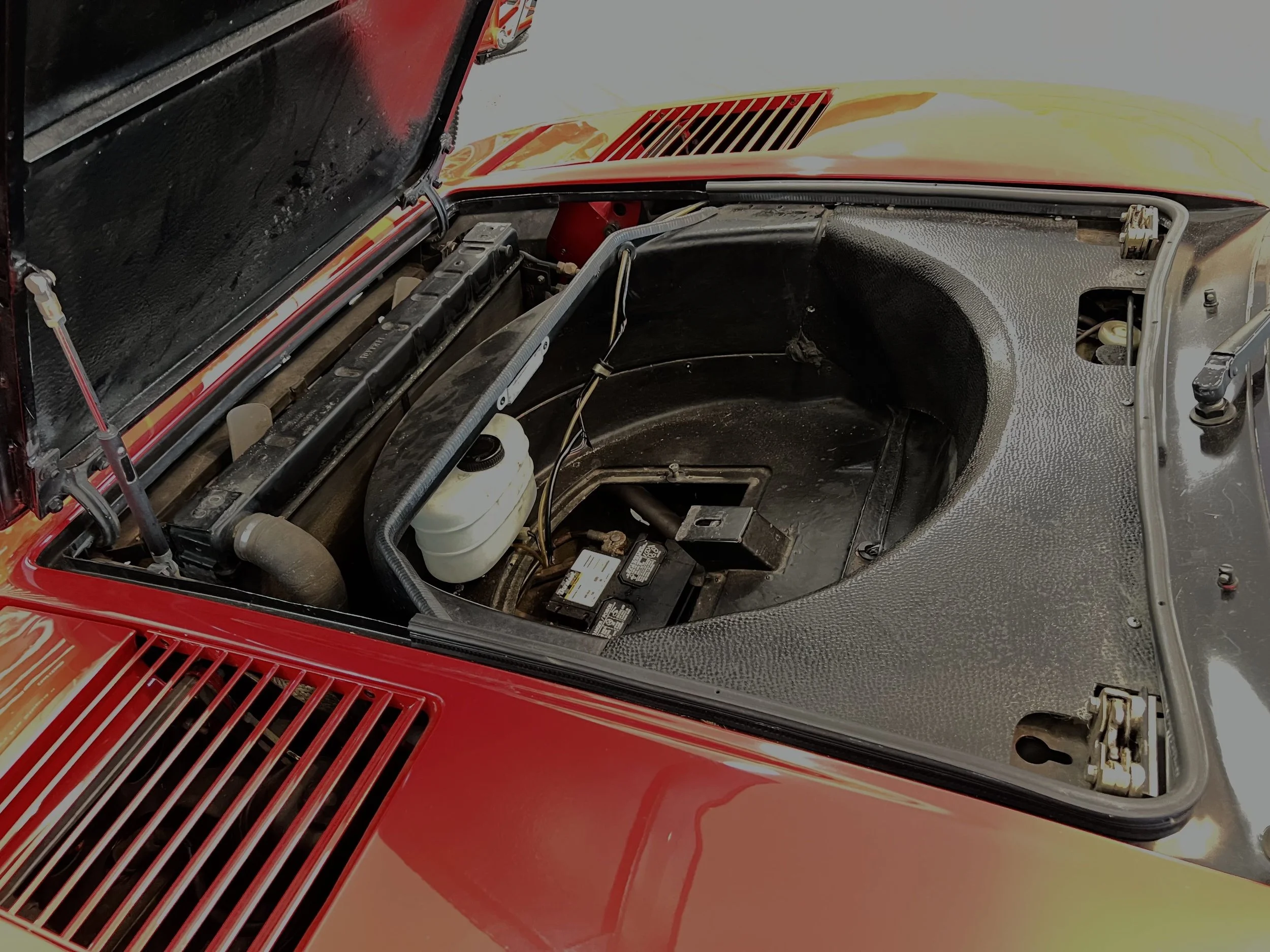 The engine bay of a red car with the hood open, showing an empty space where the engine should be, various hoses, a battery, and a coolant reservoir.