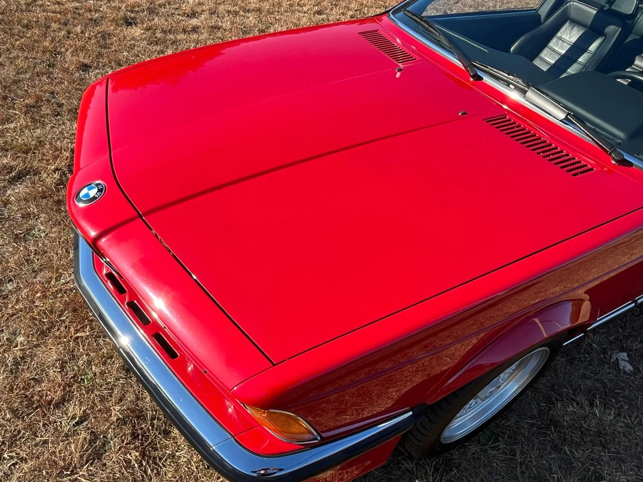 Close-up of a red BMW vintage car parked on grass, showing the front part of the hood, windshield, and interior with black seats.