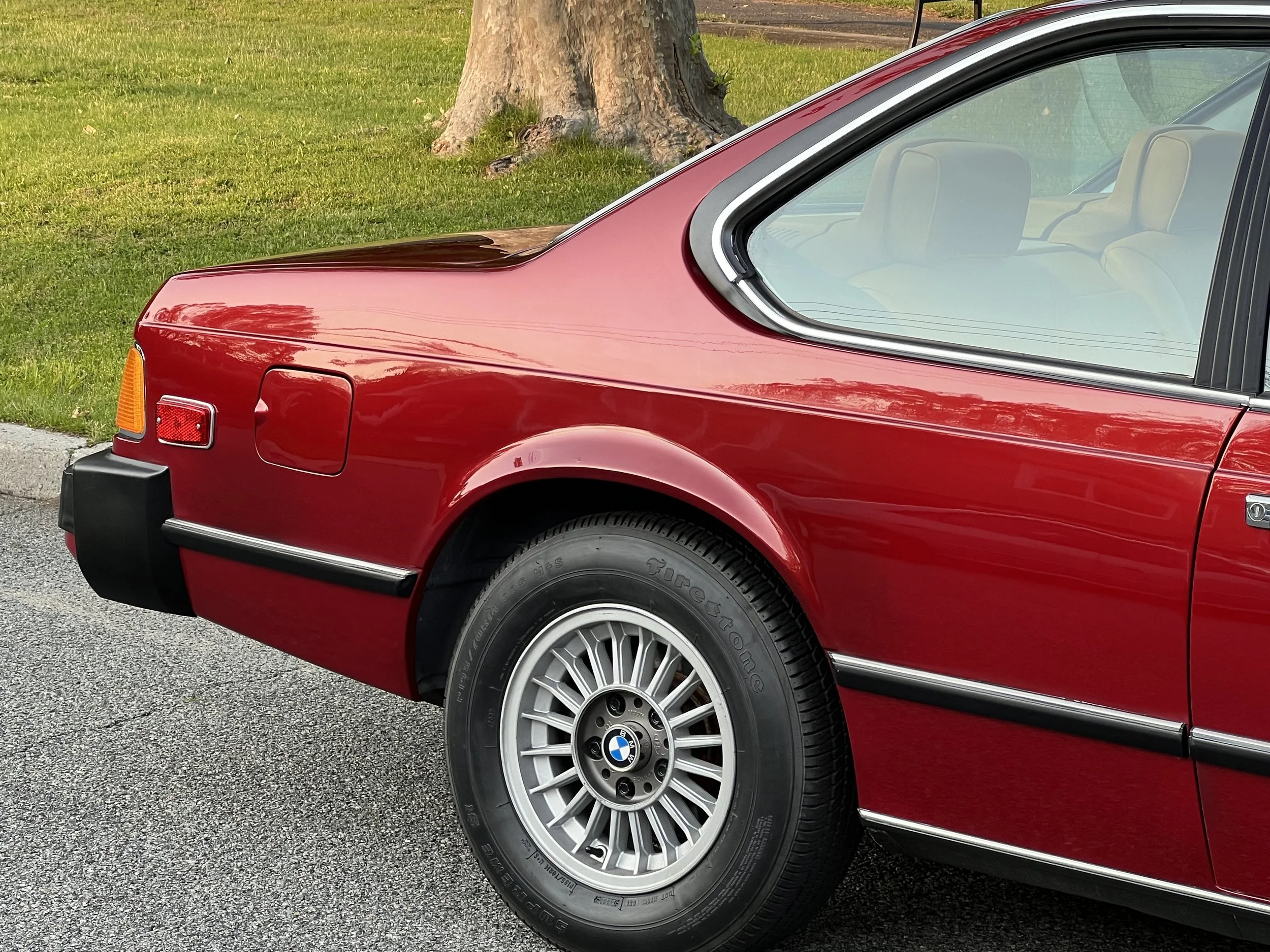 Close-up of the rear quarter of a vintage red BMW BMW 630 csi with a BMW emblem on the wheel, parked on a gray asphalt road next to a grassy area with a tree trunk.