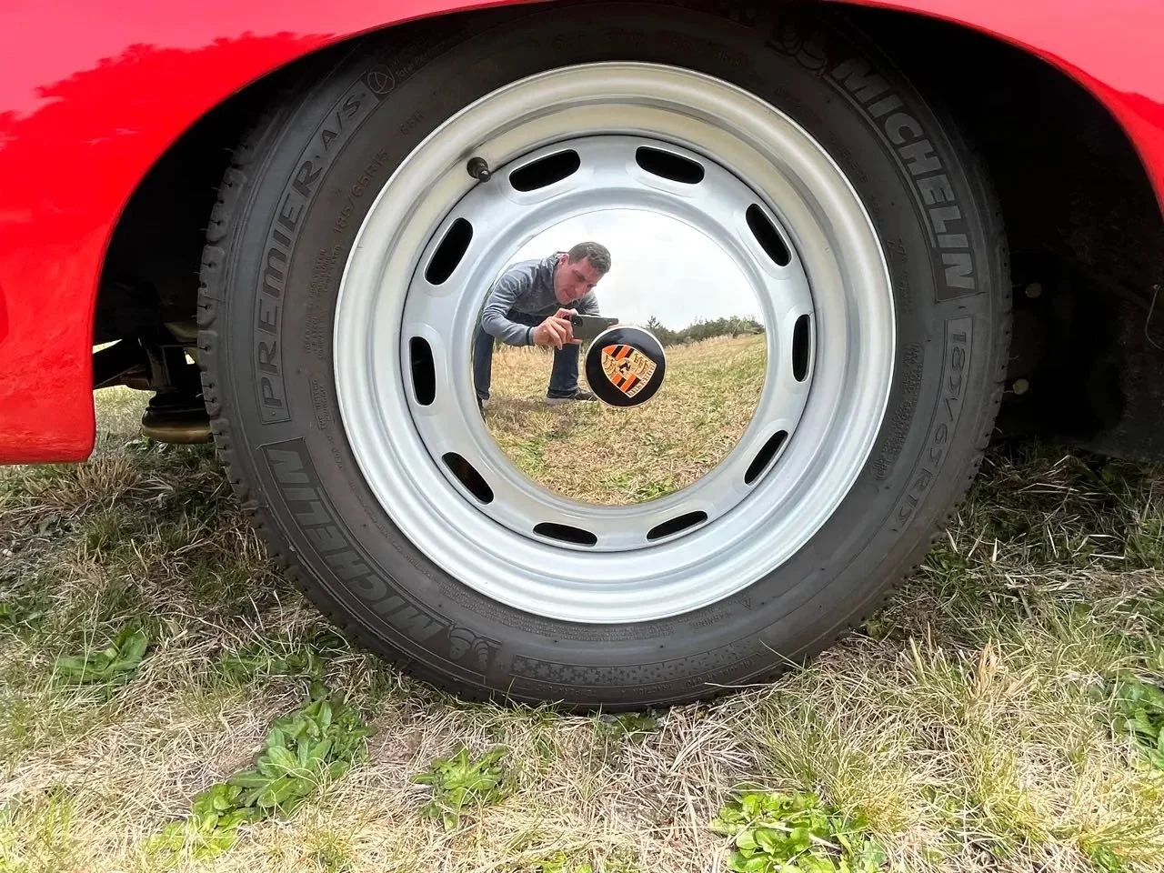 Reflection of a person taking a photo with a smartphone in the shiny surface of a car wheel, which features a Porsche logo at its center, on a grassy field.