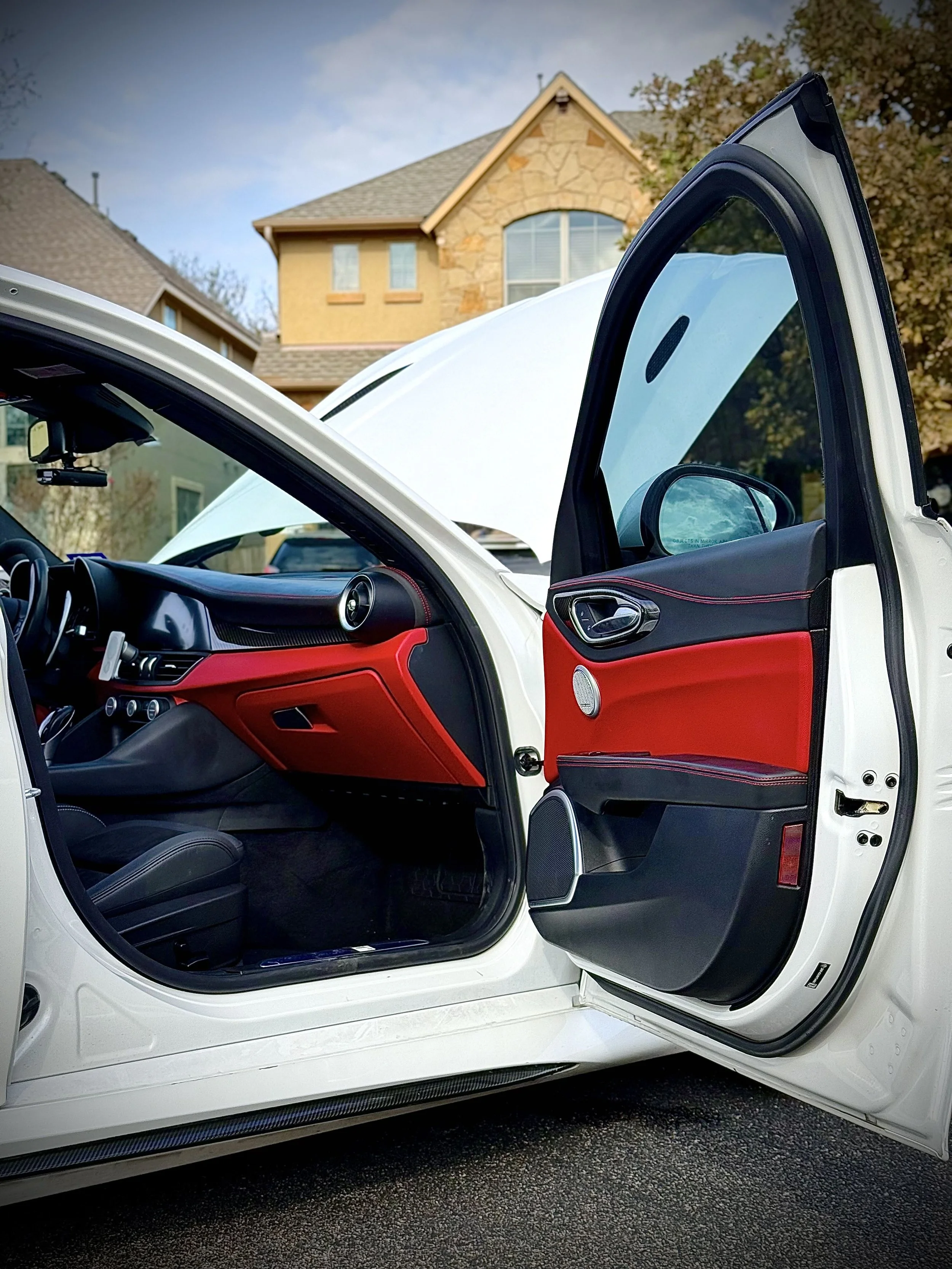Open car door revealing the vehicle's black and red interior, with a house and trees in the background.