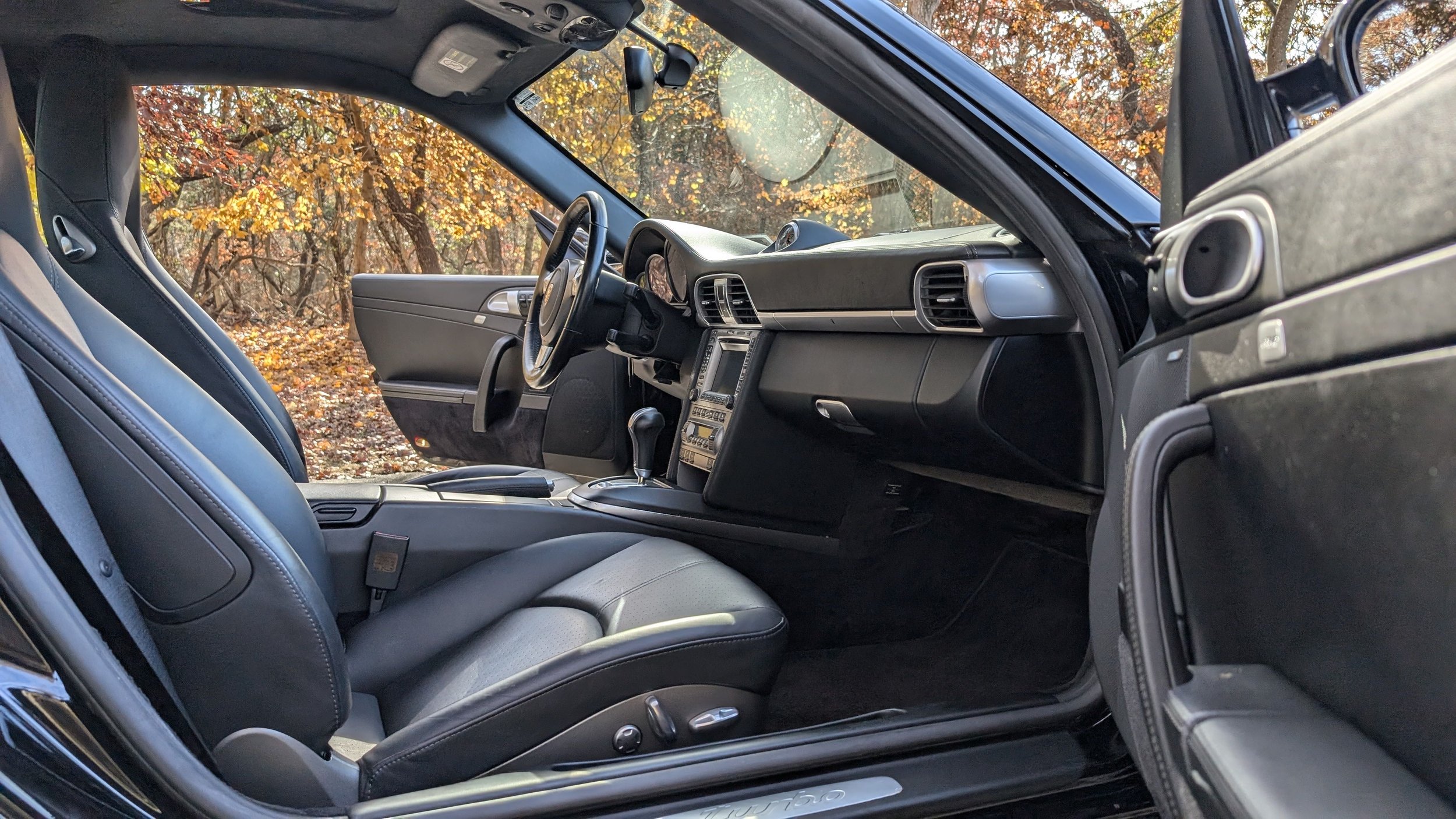 Interior of a Porsche 911 Carrera showing the front seats, dashboard, and center console, with an autumnal forest background through the windows.