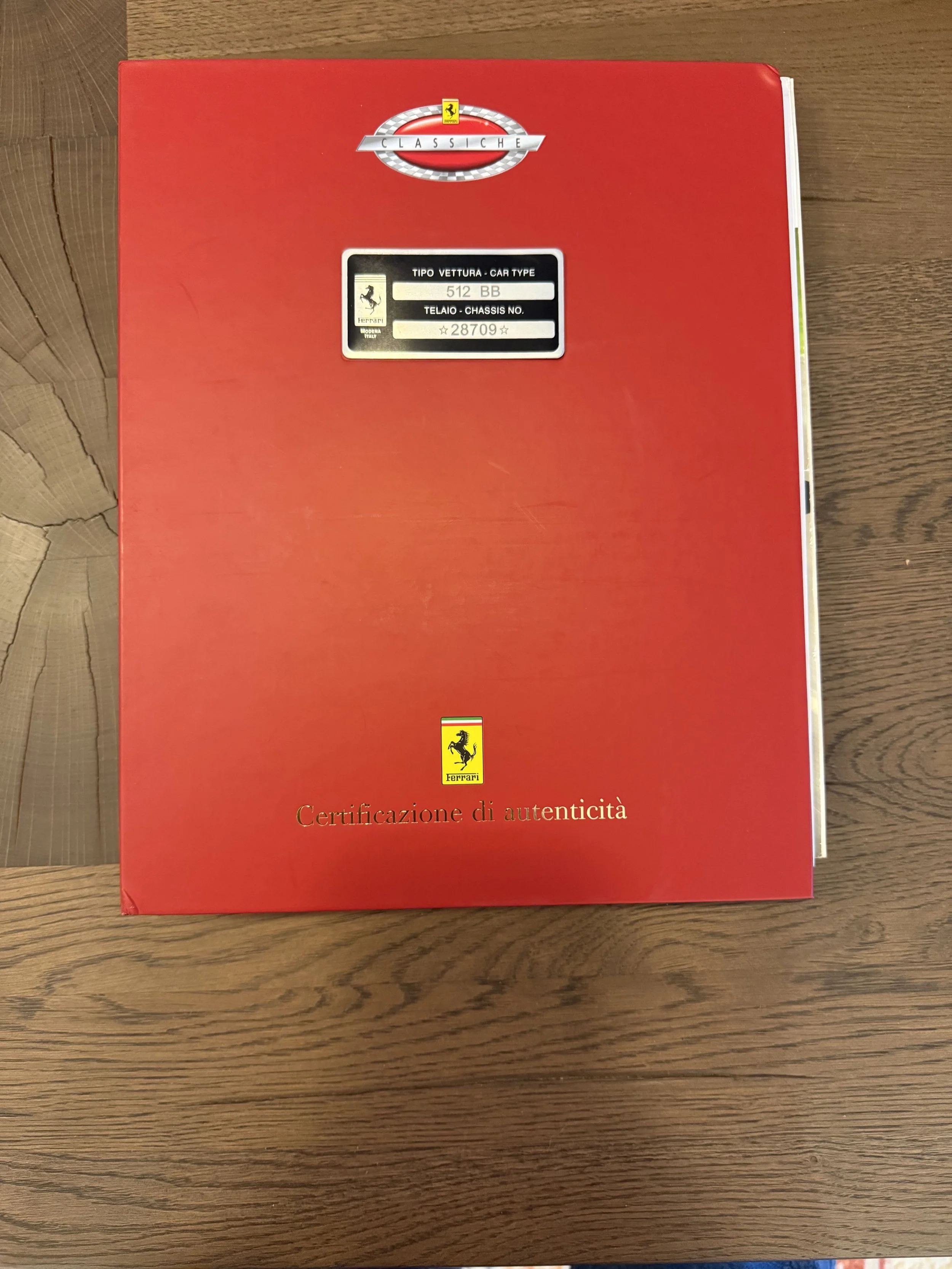 Ferrari Classiche Red Book with Ferrari branding and Italian text, placed on a wooden surface.