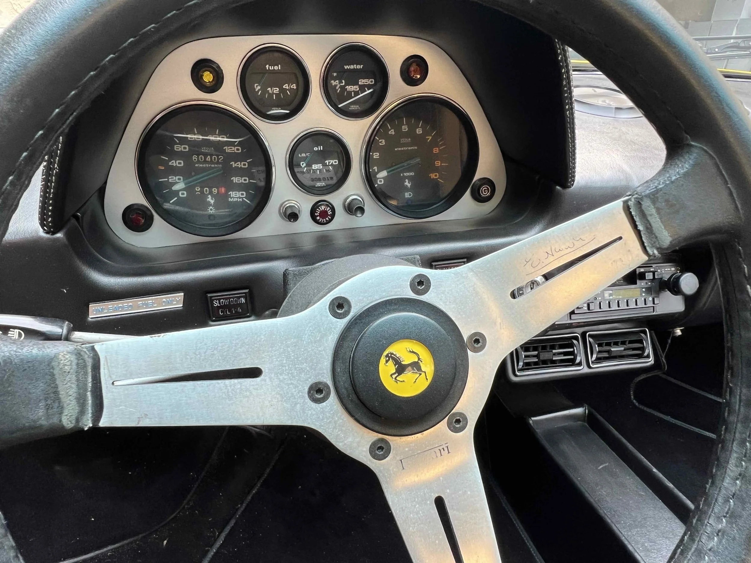 The dashboard of a vintage Ferrari sports car, showing speedometer, tachometer, and other gauges, with a signature on the steering wheel.