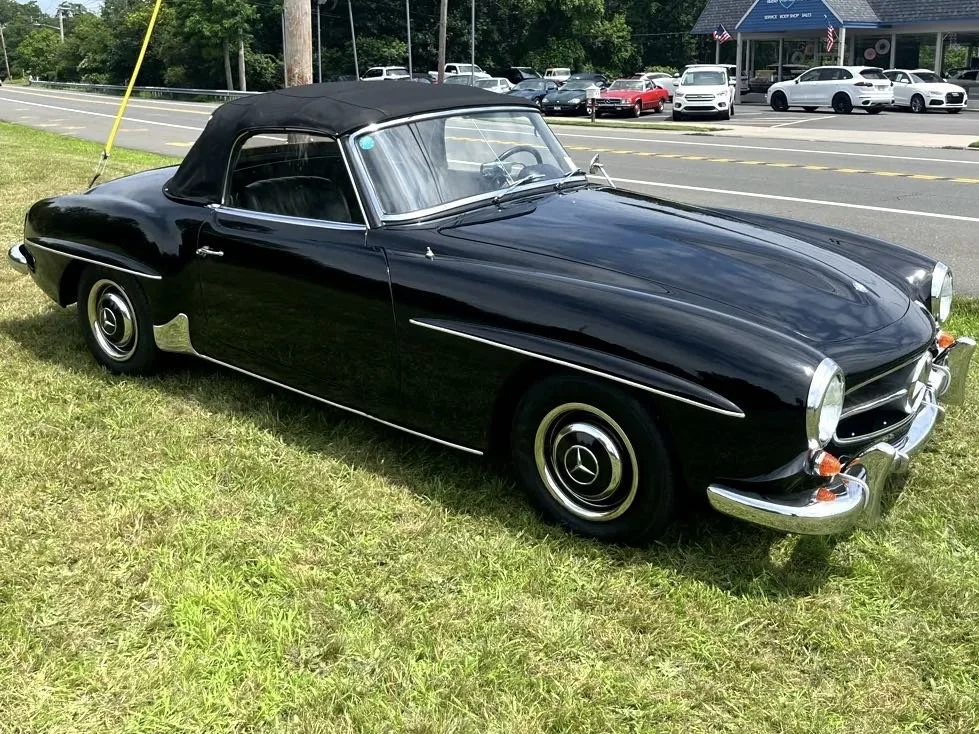 A vintage black Mercedes-Benz convertible car parked on a grassy area near a busy street with multiple cars and a building in the background.