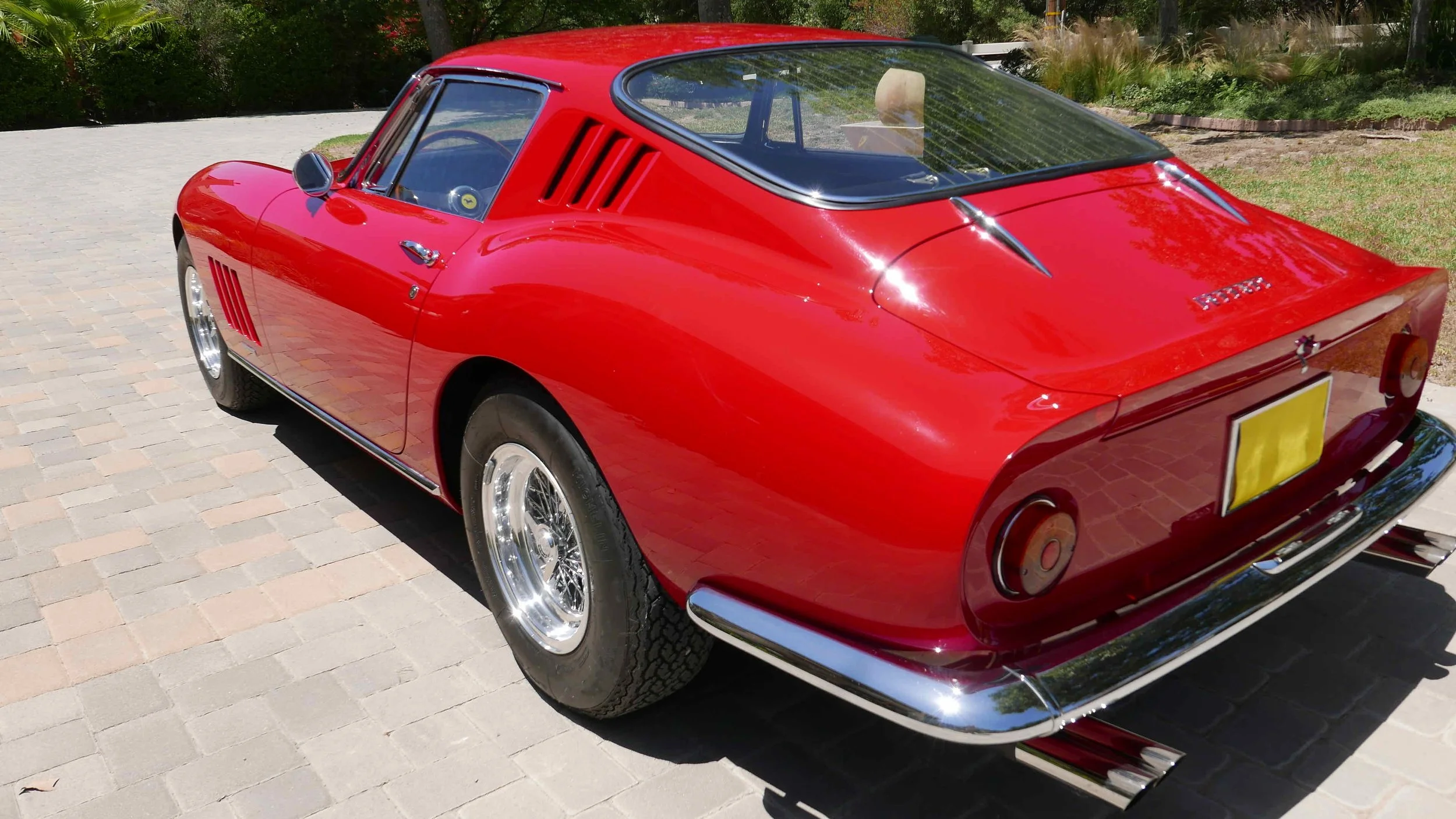 Red vintage sports car parked on a brick driveway with greenery in the background.