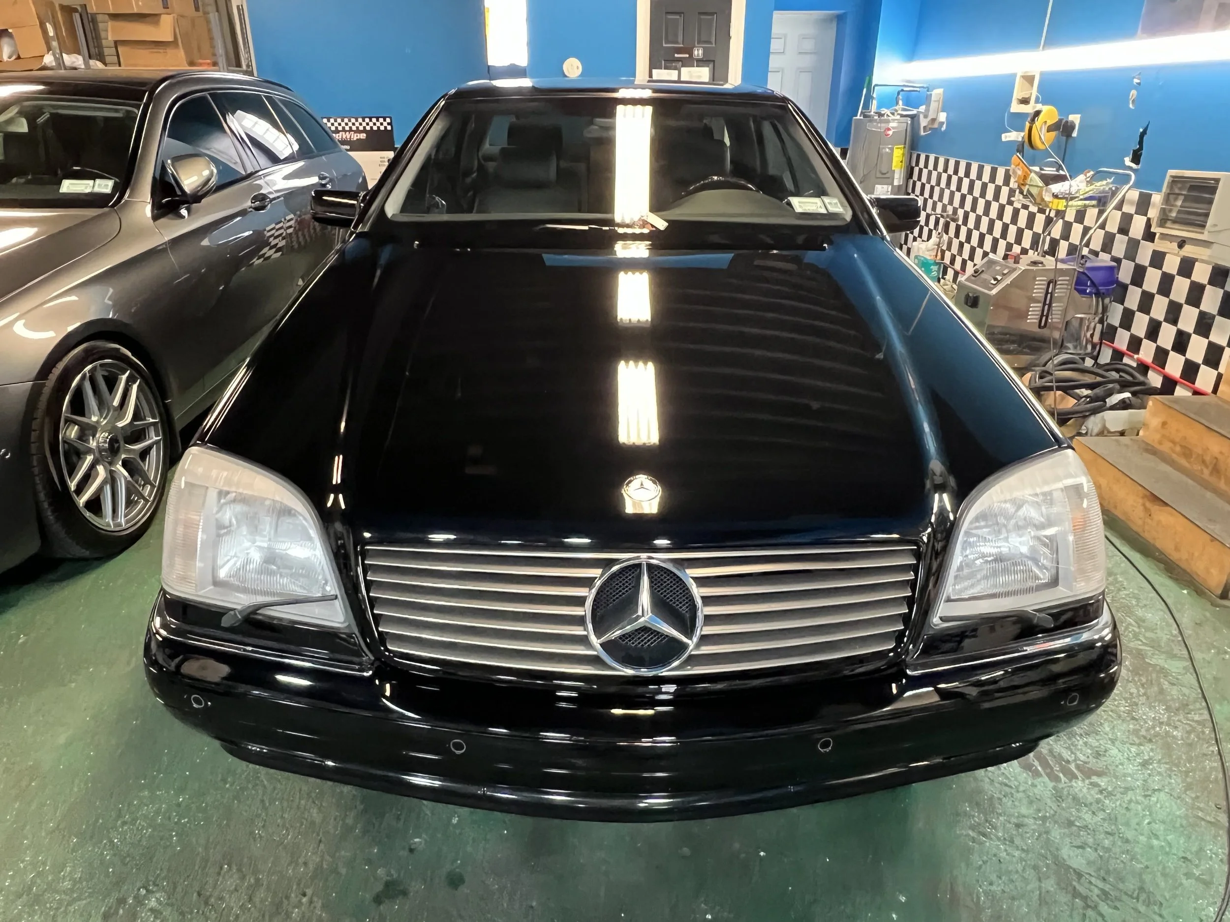 Black Mercedes-Benz car parked inside a garage with another vehicle to its left and car repair tools and equipment around.