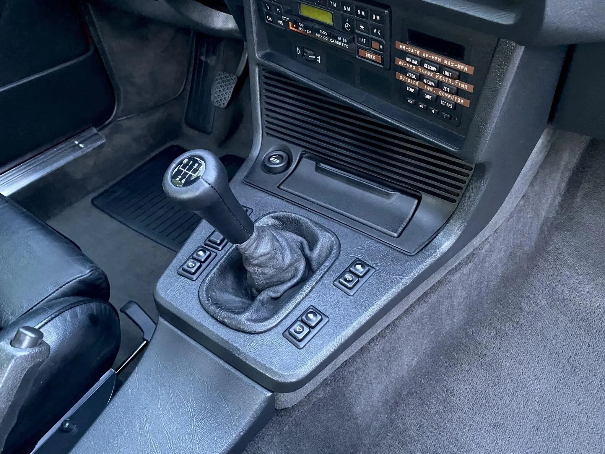Interior of a vintage car, showing the gear shift lever, dashboard, and control buttons. The gear shift is manual, with a leather boot.
