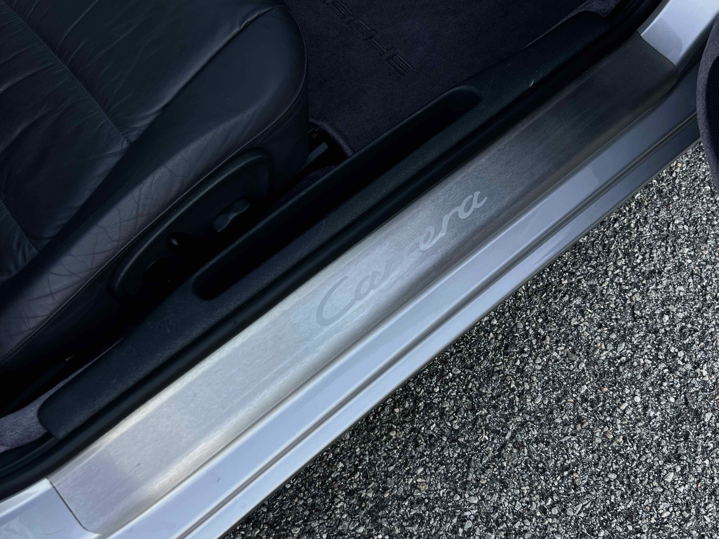 Close-up of the door sill of a Porsche Carrera with black leather seat and textured black ground surface outside.
