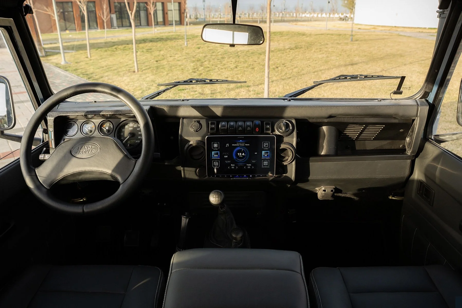 Interior of a Land Rover vehicle showing the dashboard, steering wheel, gear shift, and a touchscreen display, with a grassy area and trees visible through the windshield.