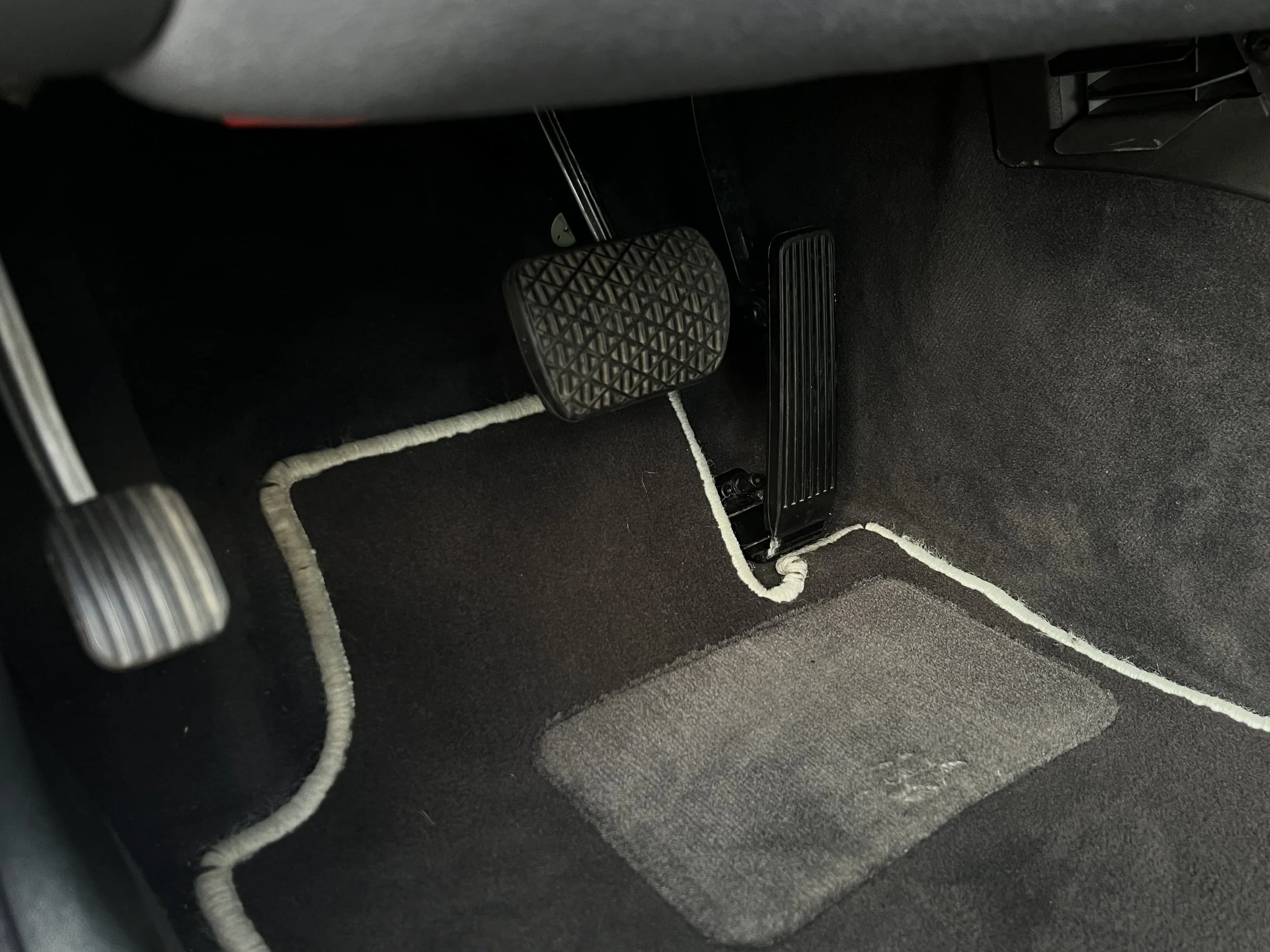 The image shows the interior of a car's footwell area, featuring the brake pedal, accelerator pedal, and a portion of the carpeted floor with white piping along the edges.