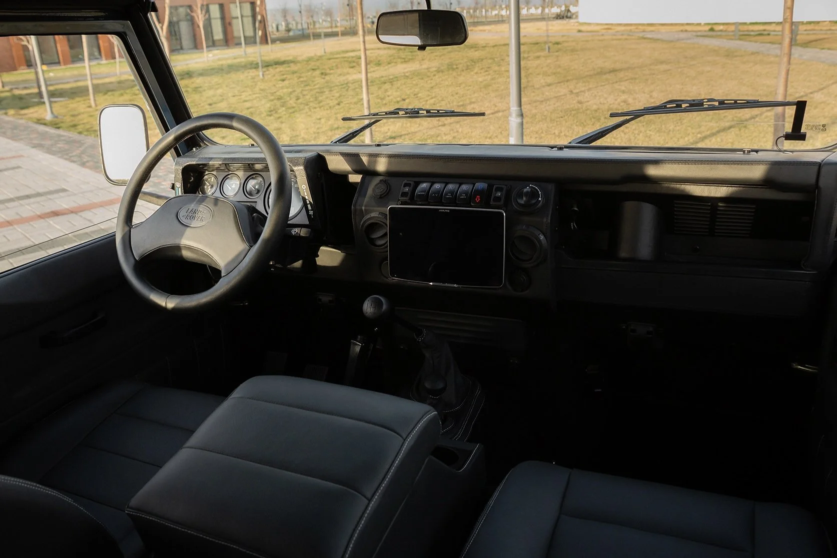 Interior of a Land Rover vehicle, showing the black dashboard with a digital screen, steering wheel, gear shift, two seats, and a large windshield with a view of a grassy area, a sidewalk, and some streetlights.