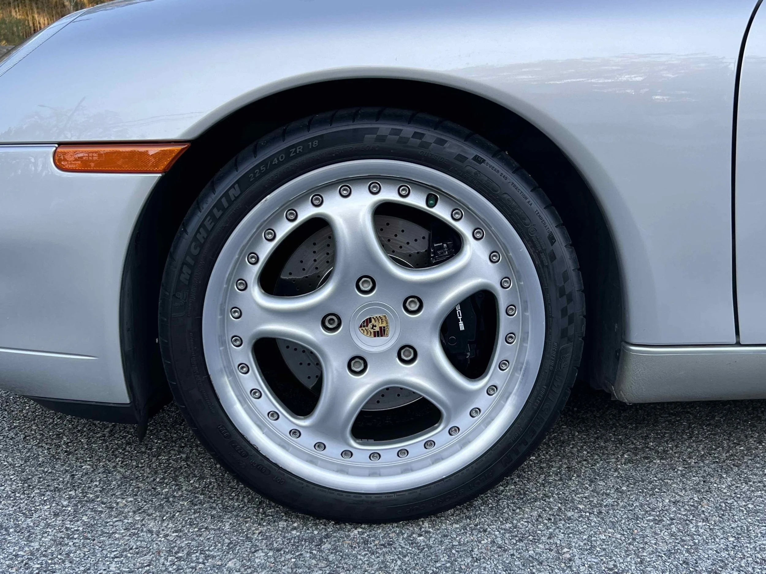 Close-up of a silver sports car wheel with a Porsche emblem, Michelin tire, and drilled brake disc, parked on a paved surface.
