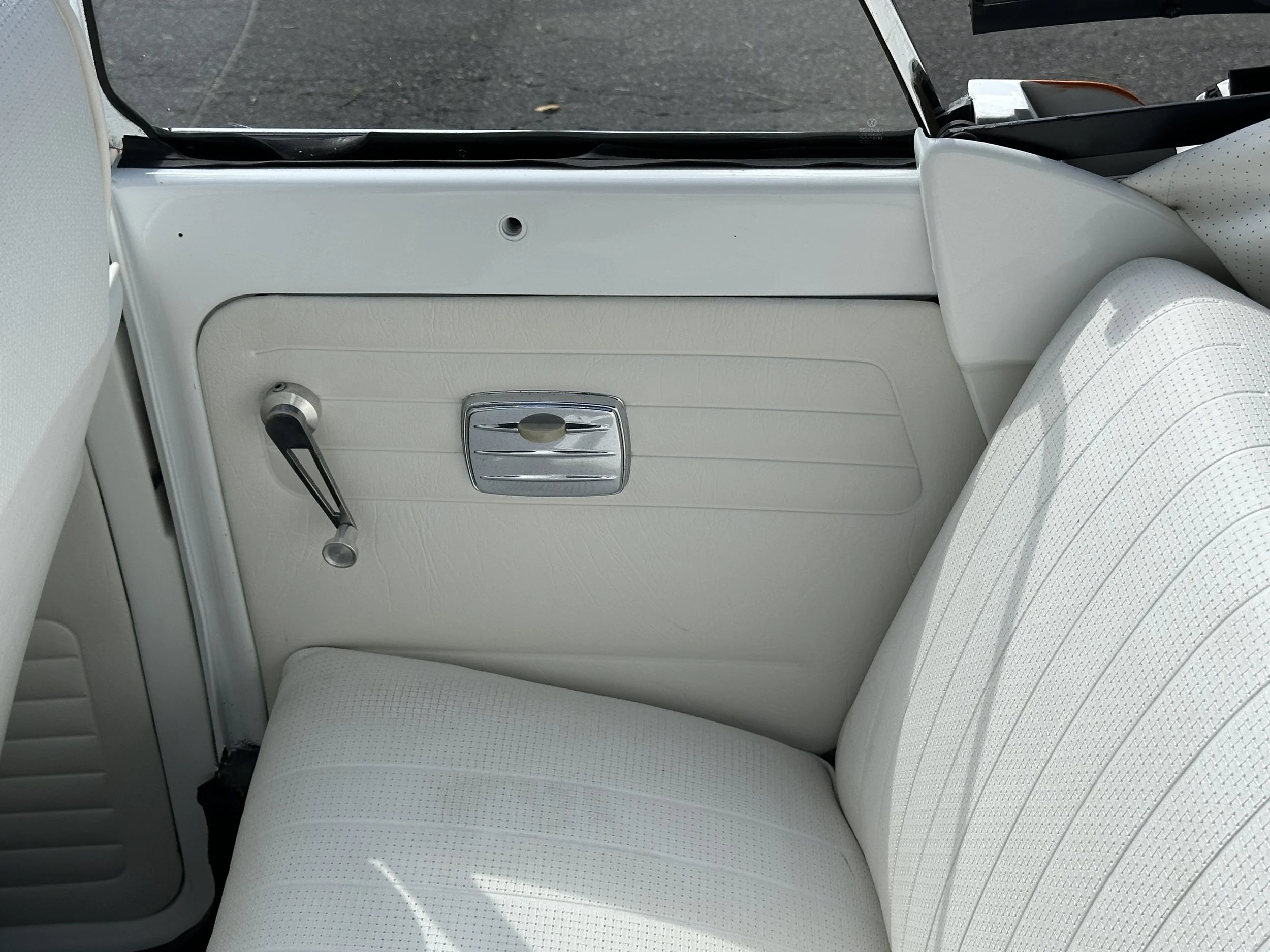 Interior of a vintage car showing the cream-colored door panel with a window crank handle, a chrome door latch, and cream seating with a dotted texture.