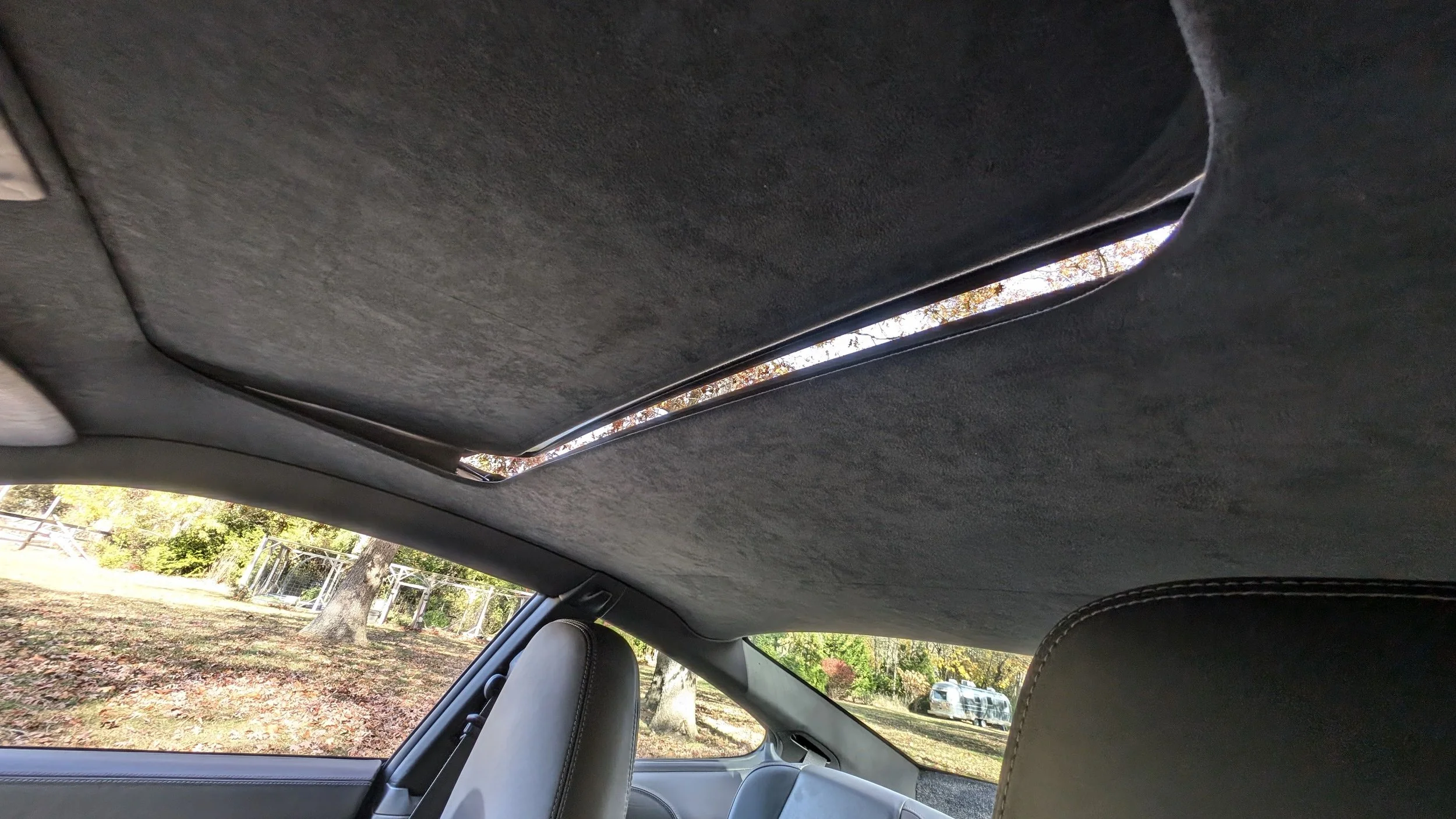 The interior ceiling of a car with a large sunroof showing an outdoor scene with trees and a fence.